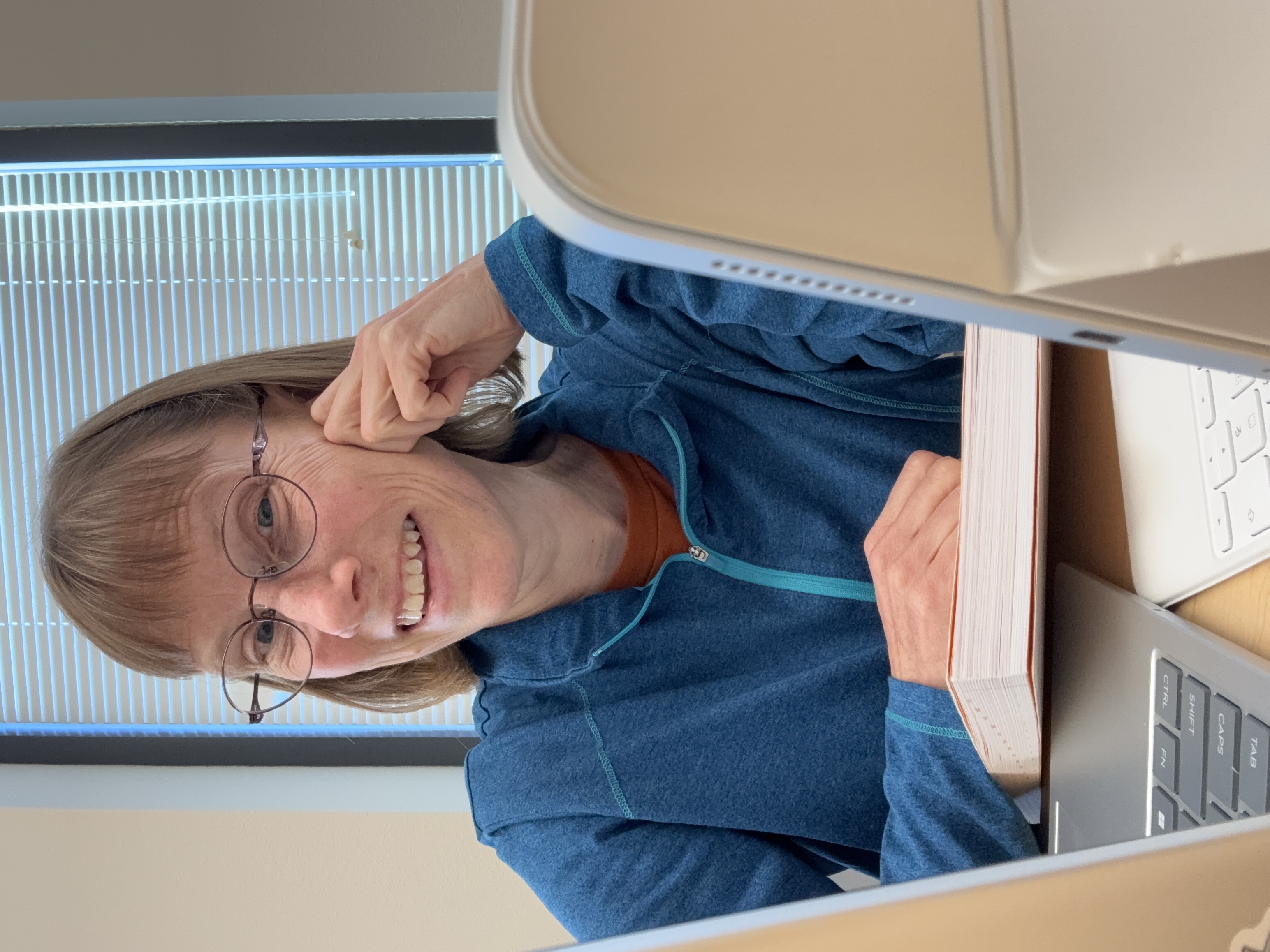 Cory smiling while holding a textbook at a desk, between a computer and an iPad.