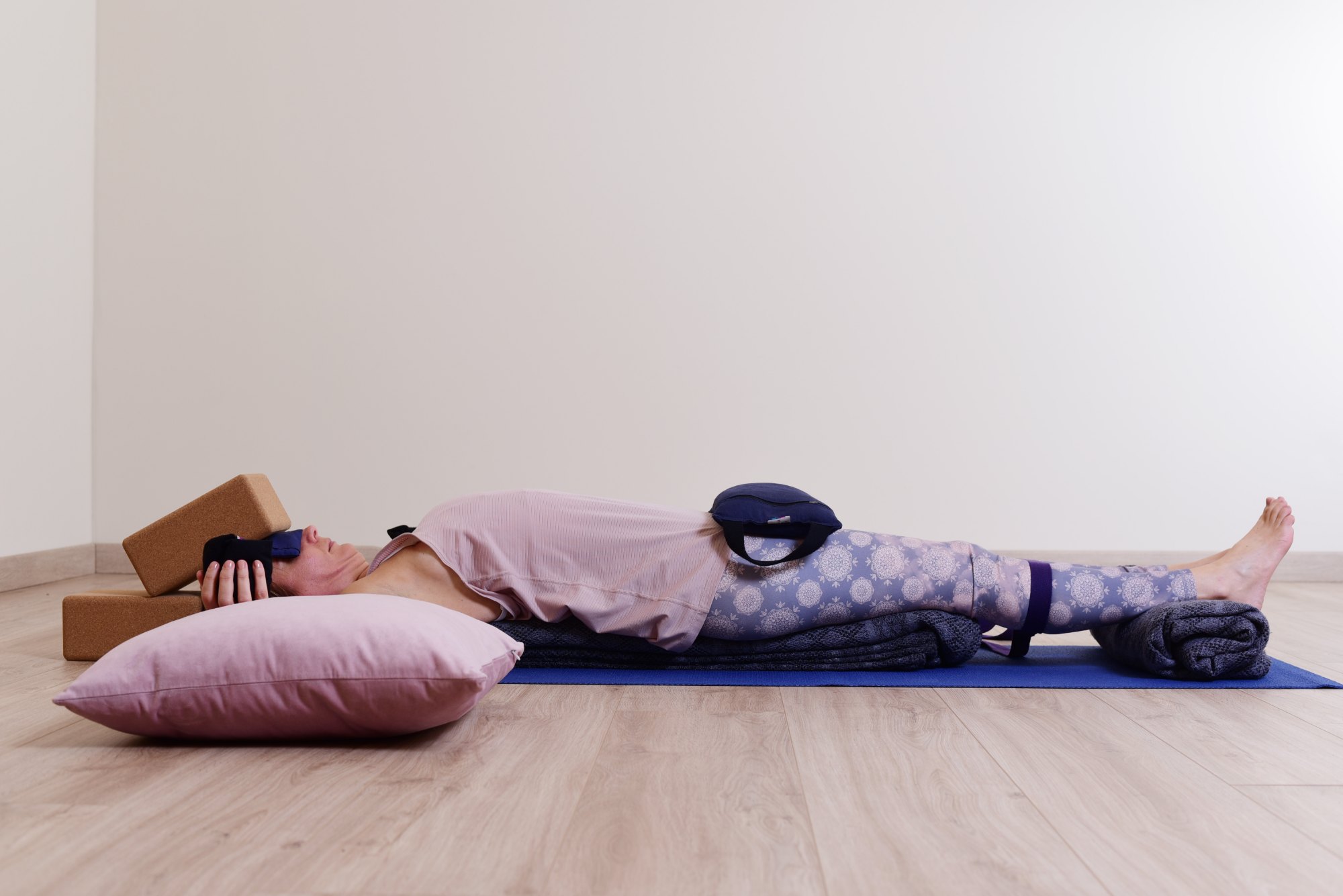 Audrey Favreau in a supported Setu Bandha Sarvangasana, a backbend from Module 2, lying over folded blankets with a strap around the thighs, a sandbag on the pelvis and blocks by the head