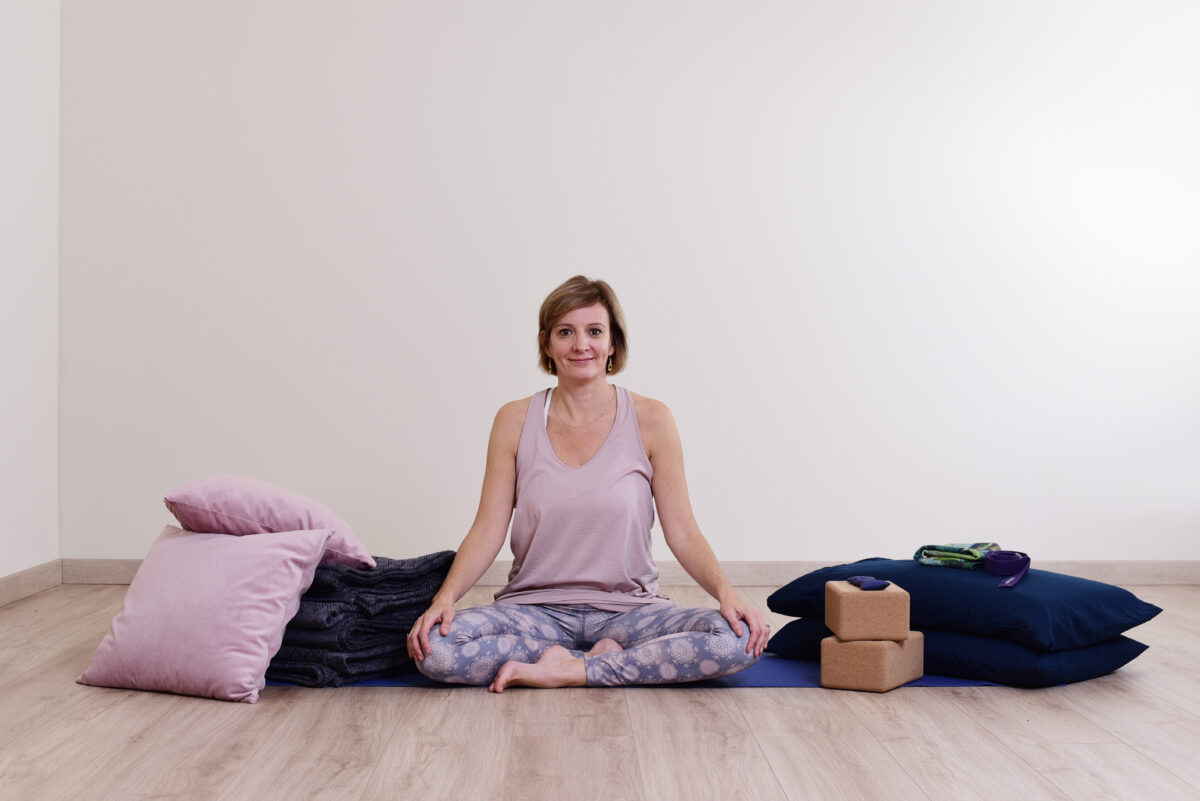 Audrey Favreau seated cross-legged on a mat in her Restorative Yoga teaching space, with pillows, blankets, bolsters and cork blocks on either side
