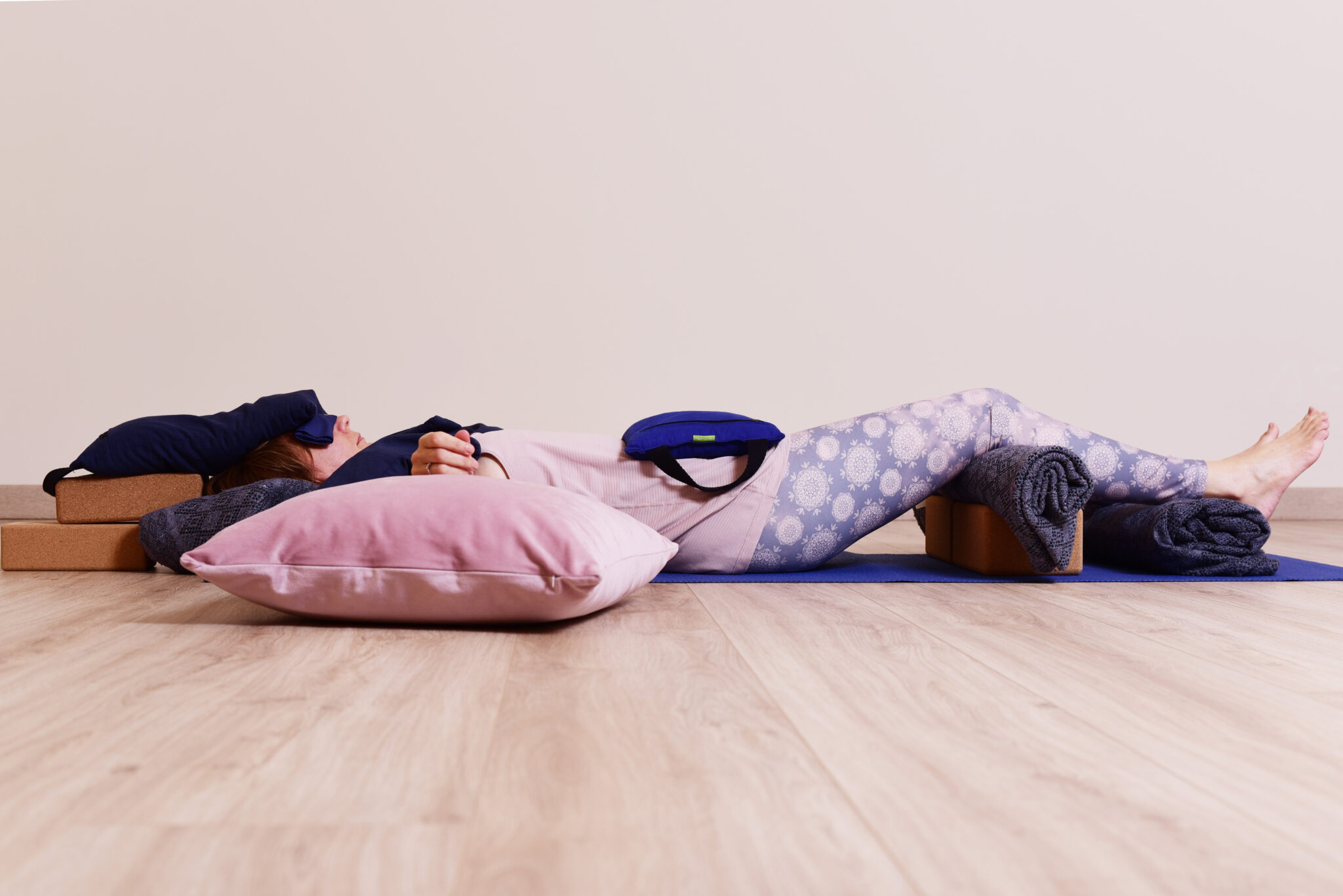 Audrey Favreau resting in a supported Savasana, head on blocks and a bolster, an eye pillow and a sandbag on the body, knees over blocks and rolled blankets