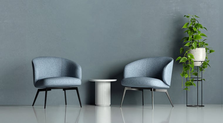 Two blue fabric armchairs facing each other with a white round table in between. A potted plant stands to the right of the chairs. The chairs and table are on a polished concrete floor against a blue wall.