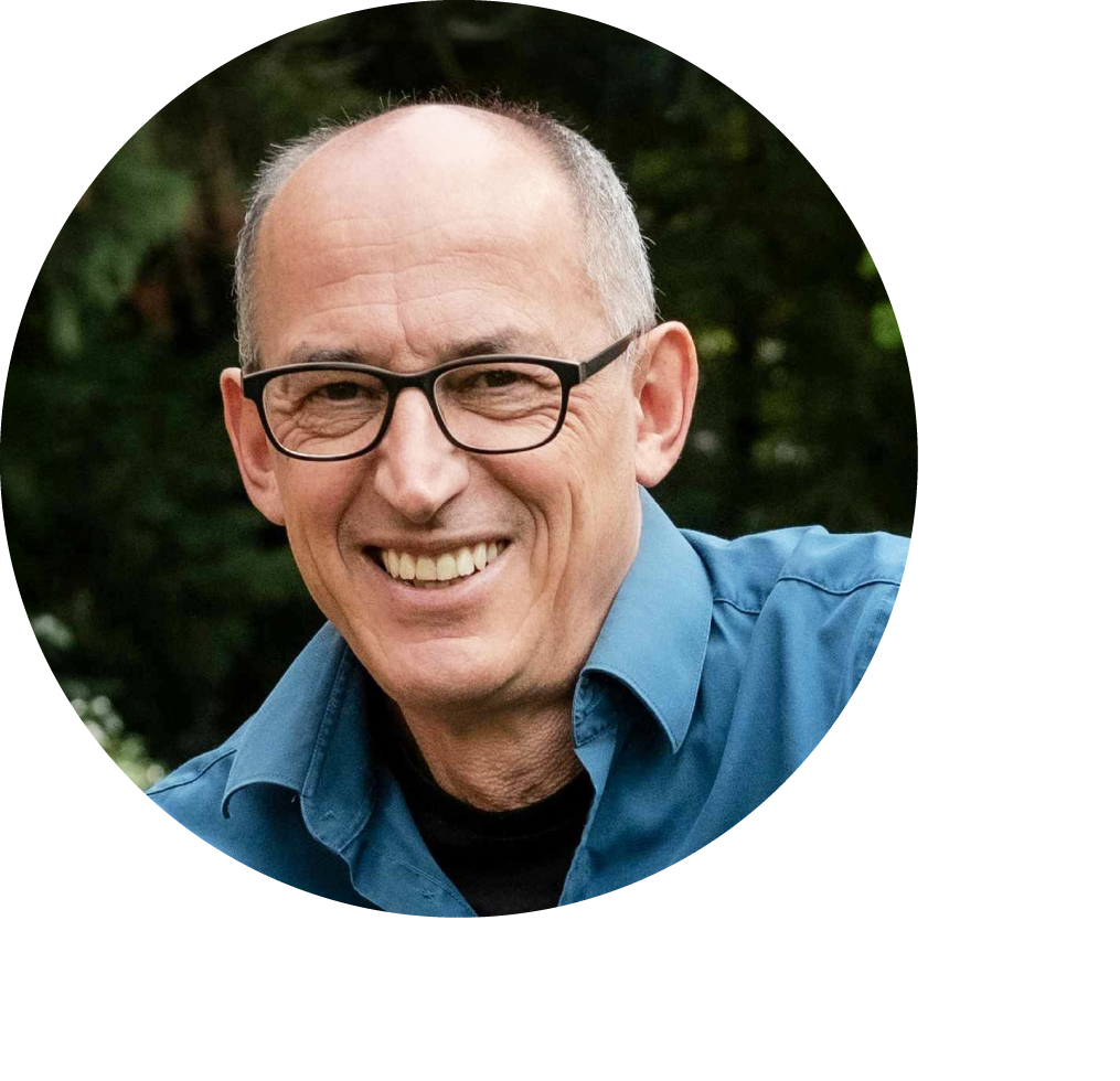 Lachender Dr. Rainer Wohlfarth mit kurzen grauen Haaren und einer schwarzen Brille vor dunkelgrünen Laubbäumen im Hintergrund