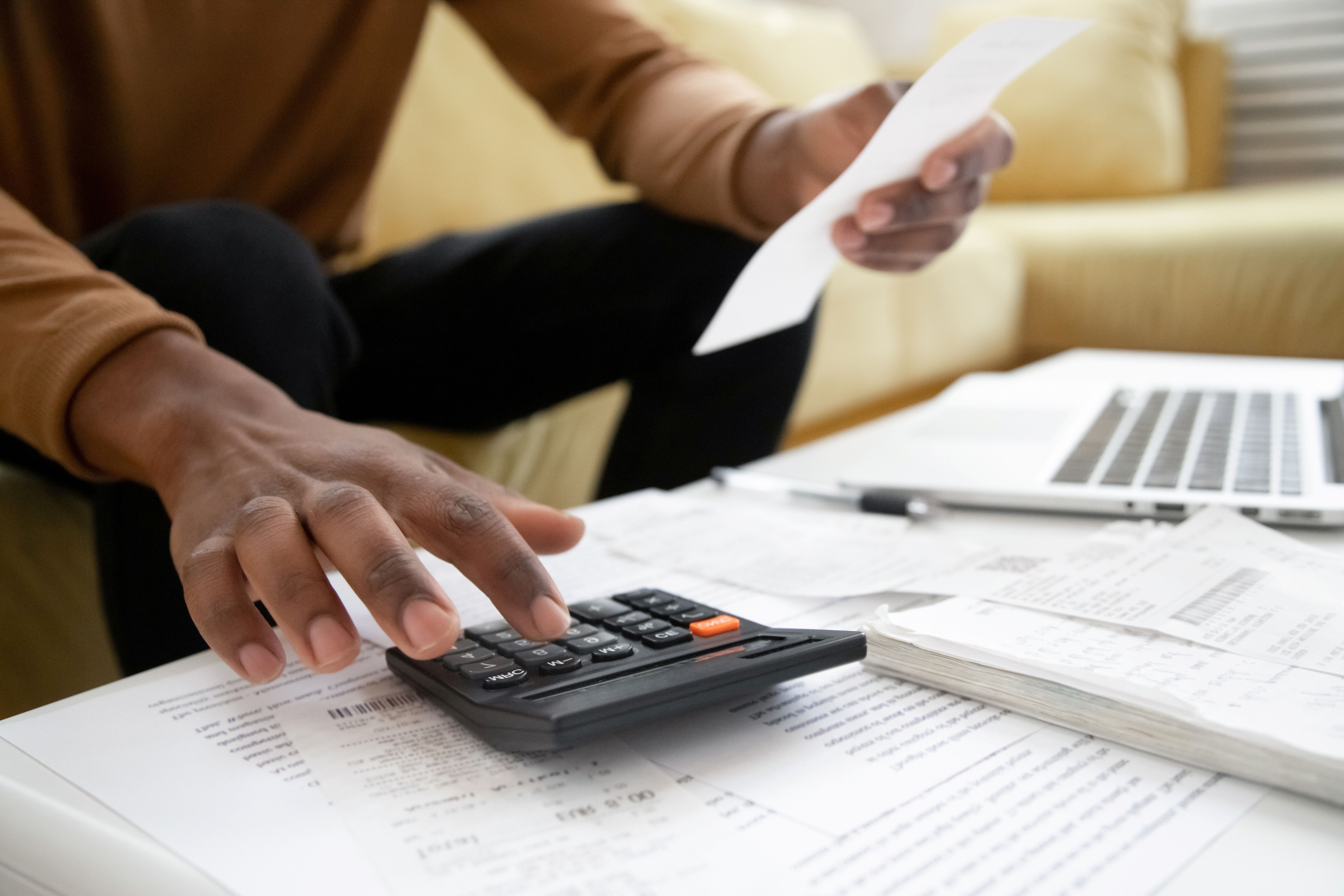 a dark skinned man sits on a pale yellow sofa with only parts of his torso, arms and legs in the picture as he leans forward.  He's holding a piece of paper in one hand and using a calculator with the other.  The calculator sits on a low table covered in paper, a notebook, pen and open laptop