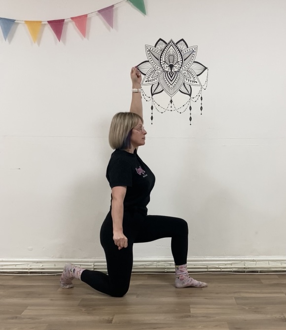 woman kneels on her right knee with her toes tucked under, her left foot on the floor in front of her and left arm up to the ceiling