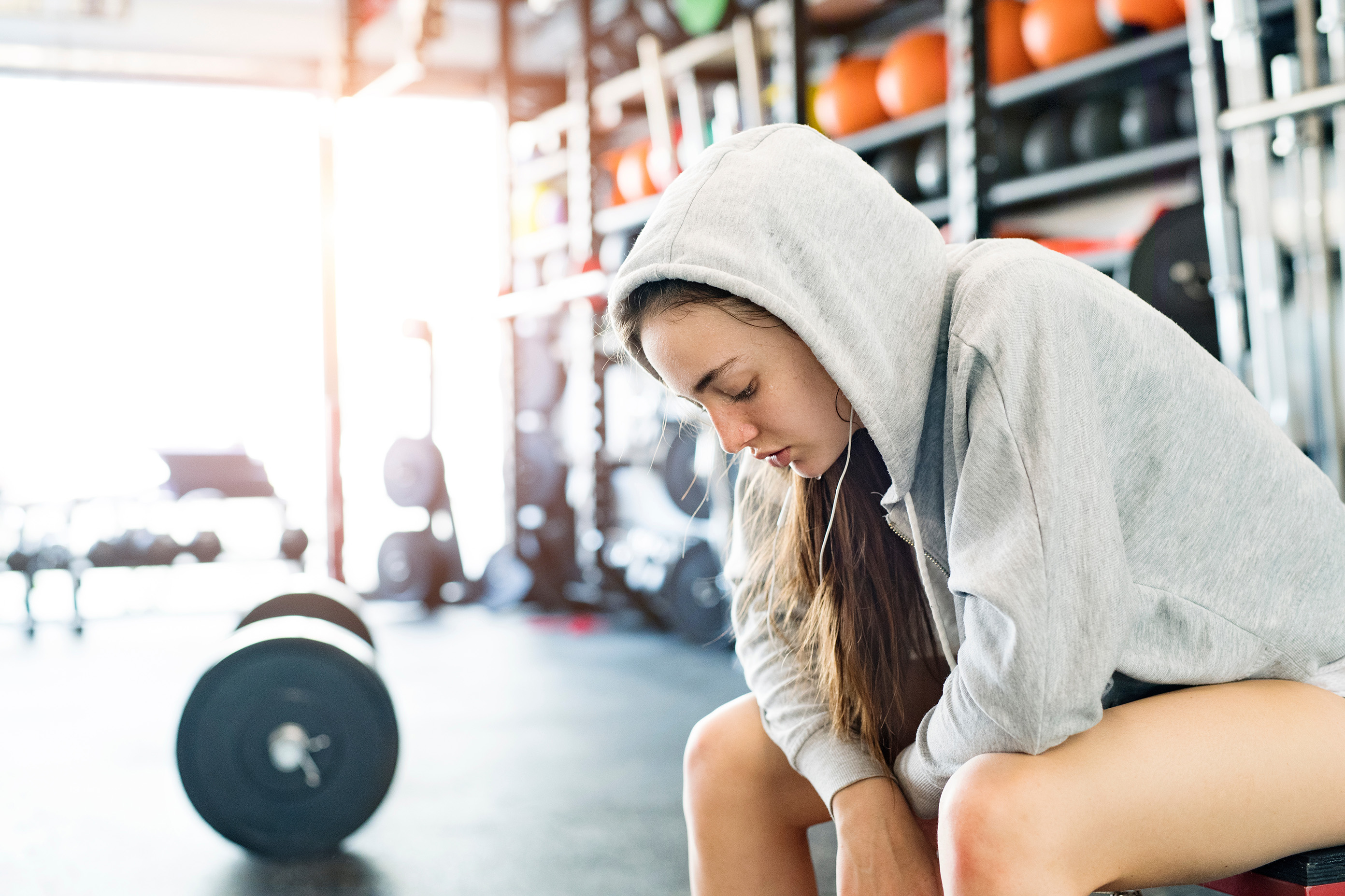 a young woman sits on a bench in a gym.  She has a hoodie on with the hood up and is leaning on her knees looking dejectedly at the floor