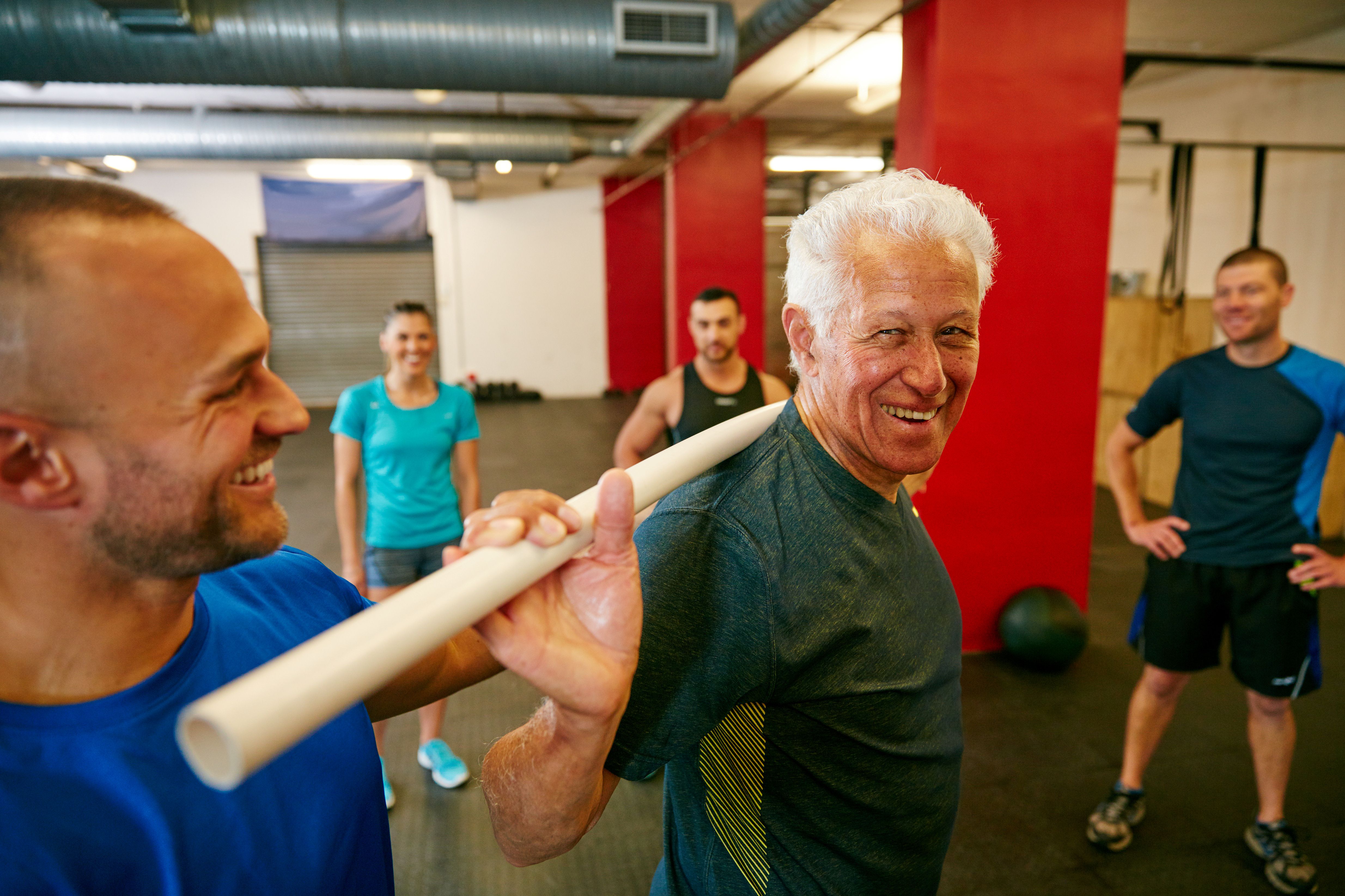 an older man has a pipe over his shoulders as a trainer helps him and other gym attendees watch