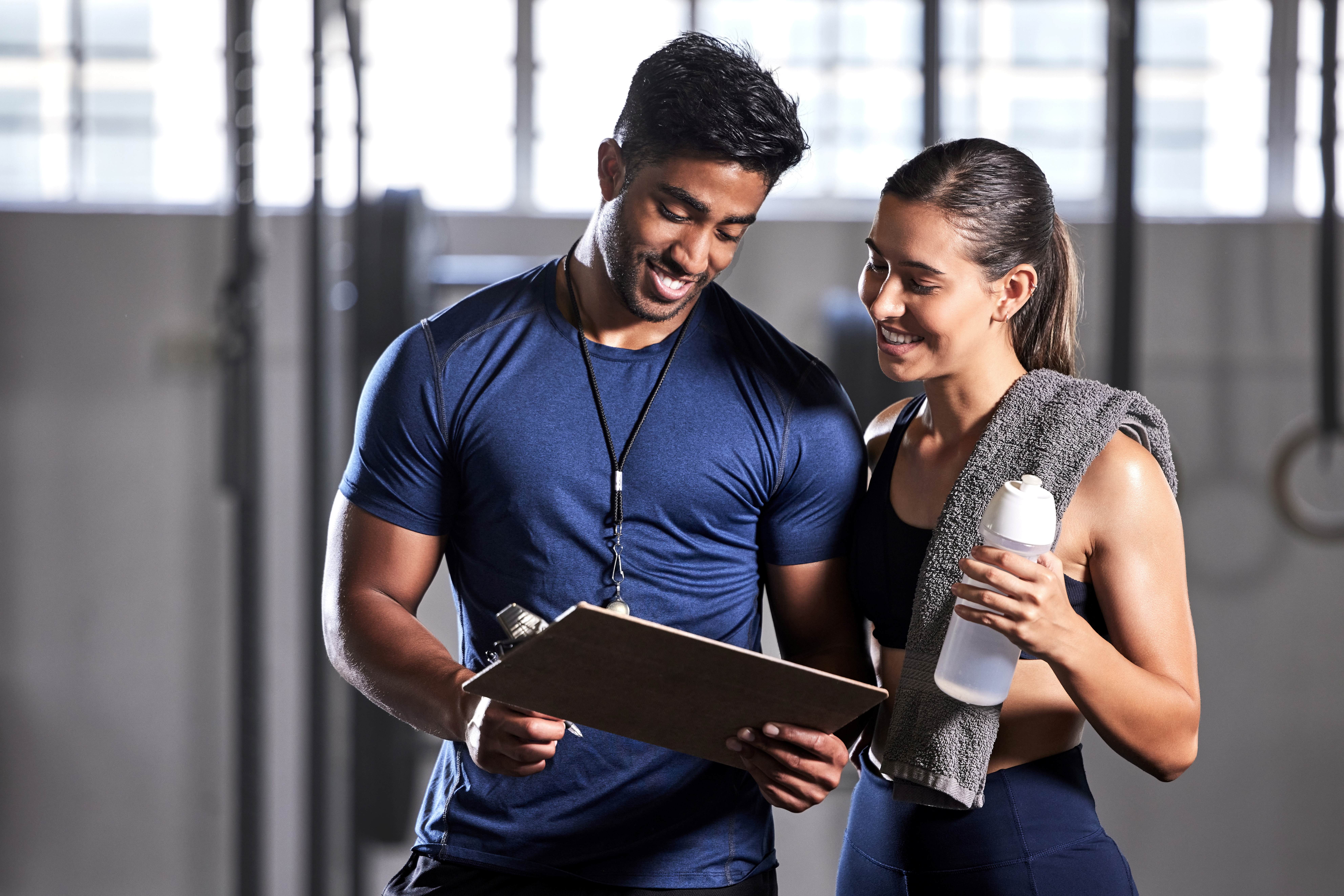 In a gym a guy with a whistle round his neck shows a woman in fitness gear soemthing on a clipboard.