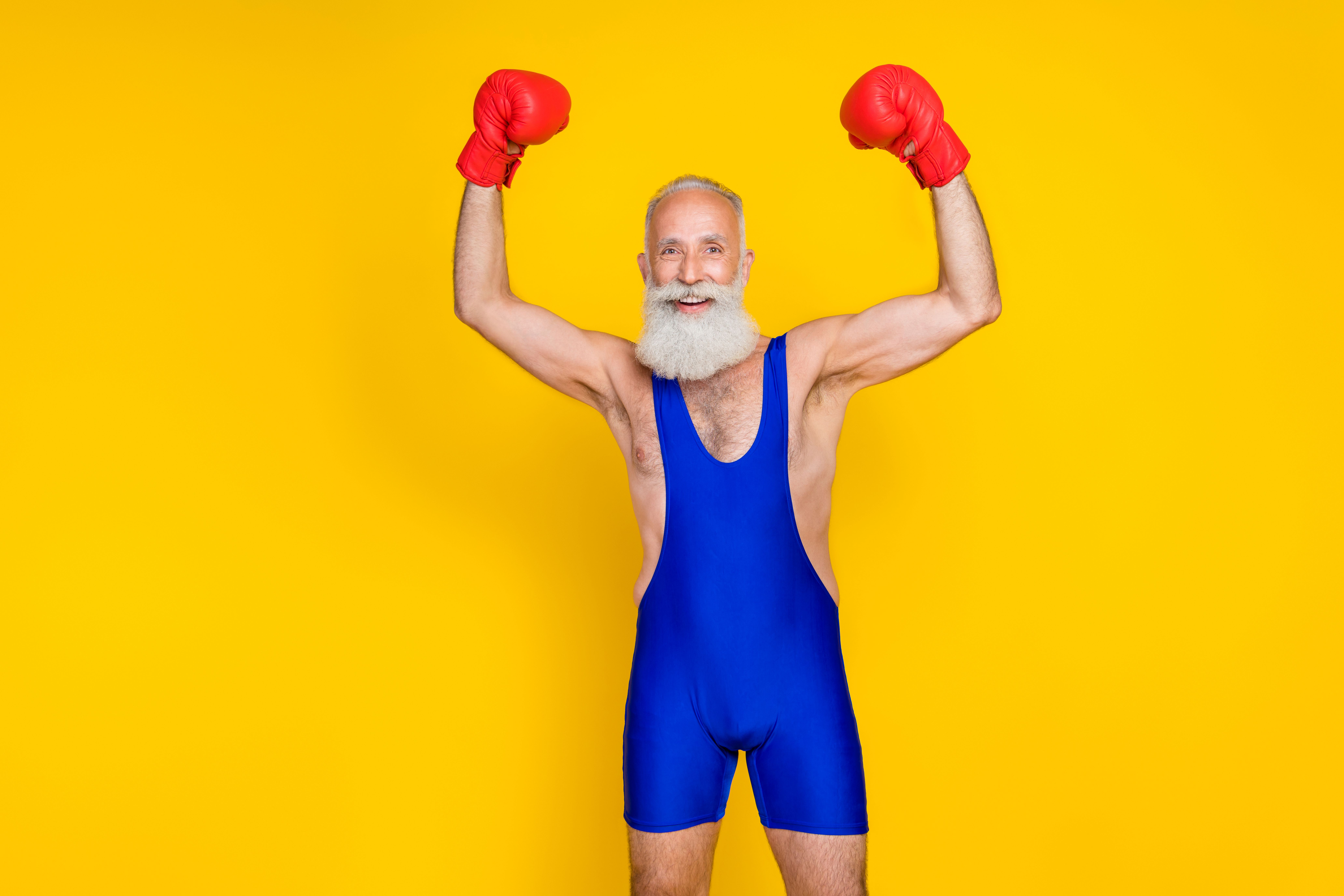 grey haired, older man with a beard wearing blue sportswear and red boxing gloves with arms up celebrating victory