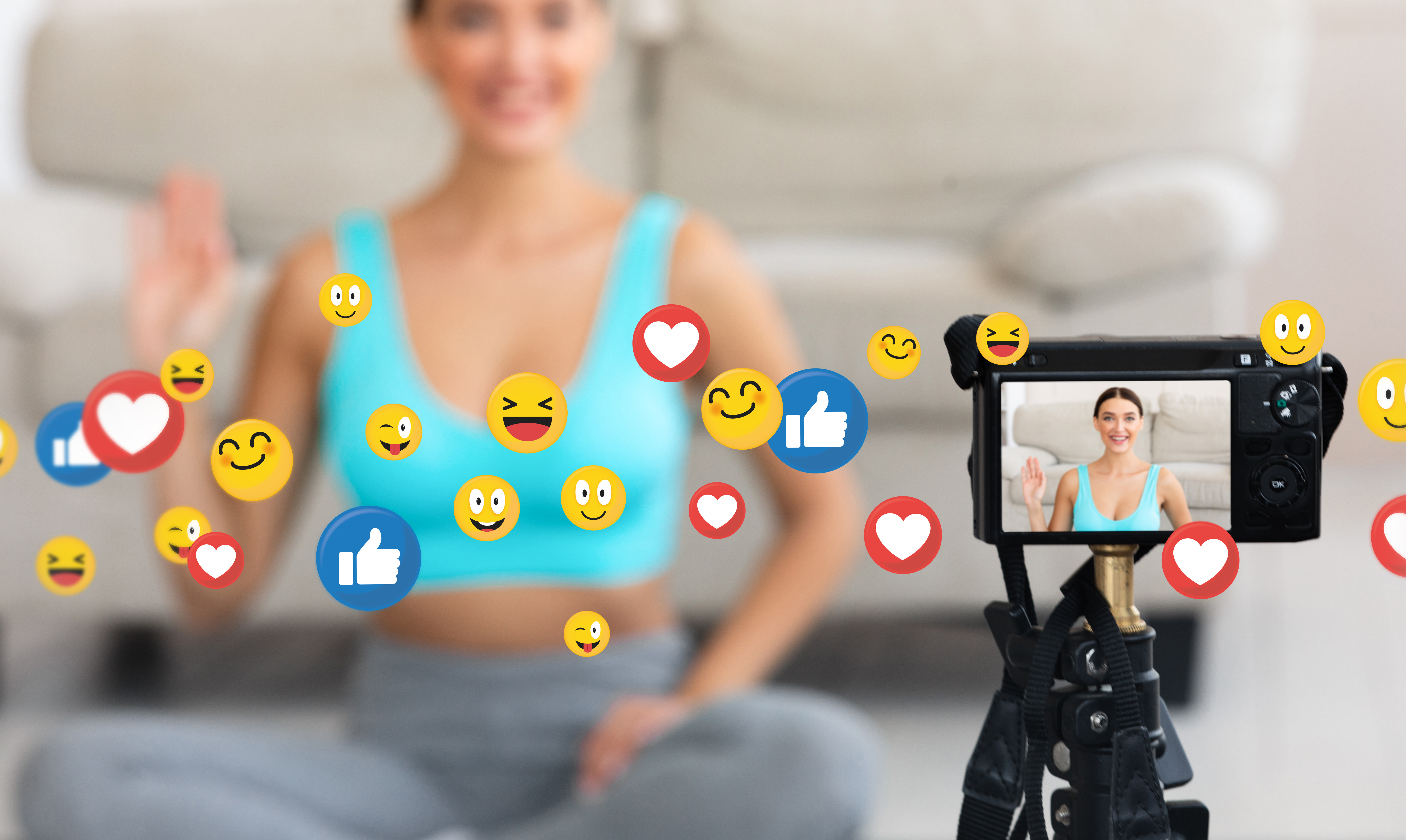 the view screen of a camera which is pointed at a woman in a crop top who is sat on the floor in a blurred background.  In front of her across the image are social media reaction emojis 