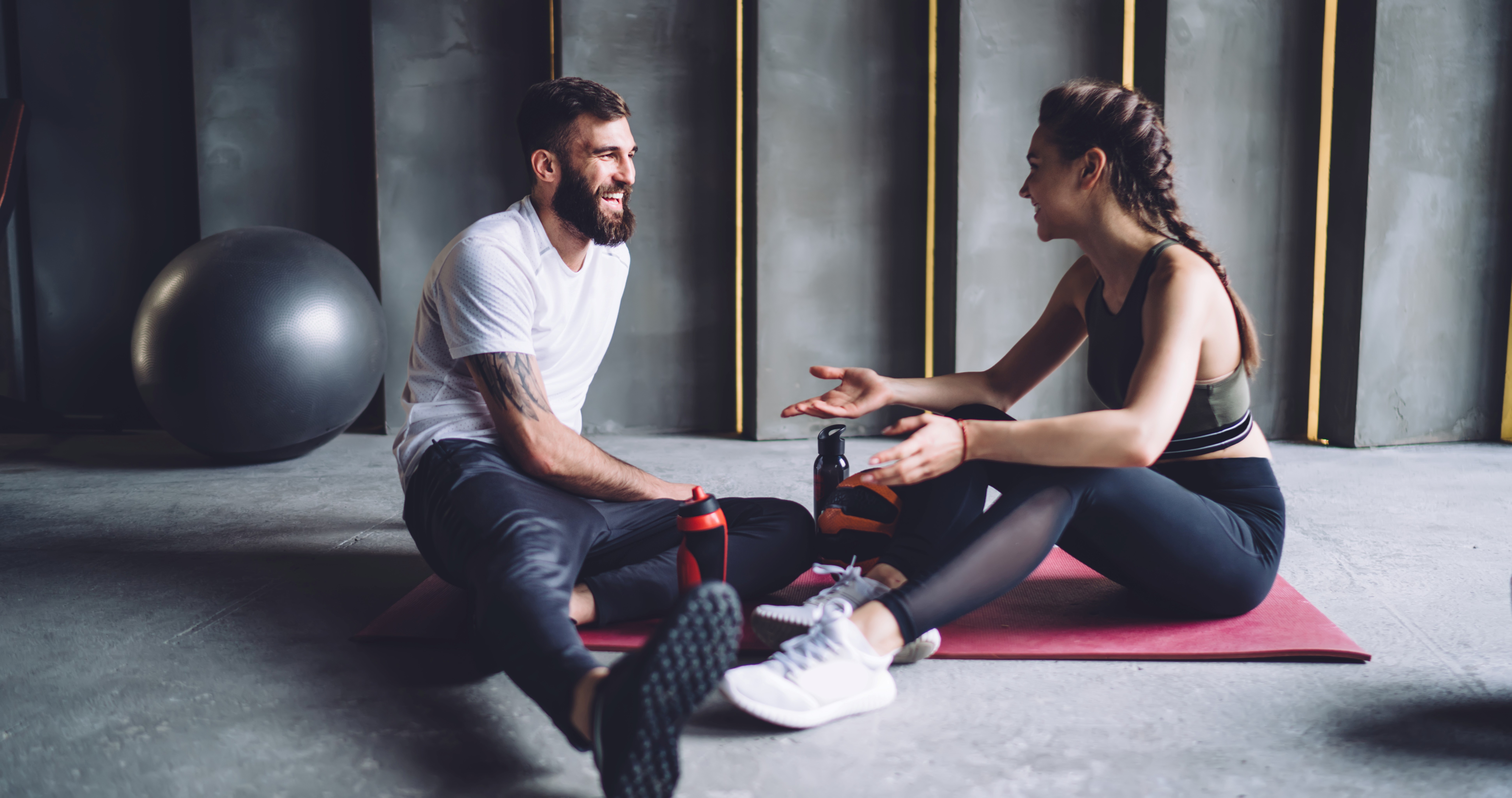a man and woman in their 20s are sat on the floor in a fitness space wearing fitness clothing.  they are smiling and chatting.