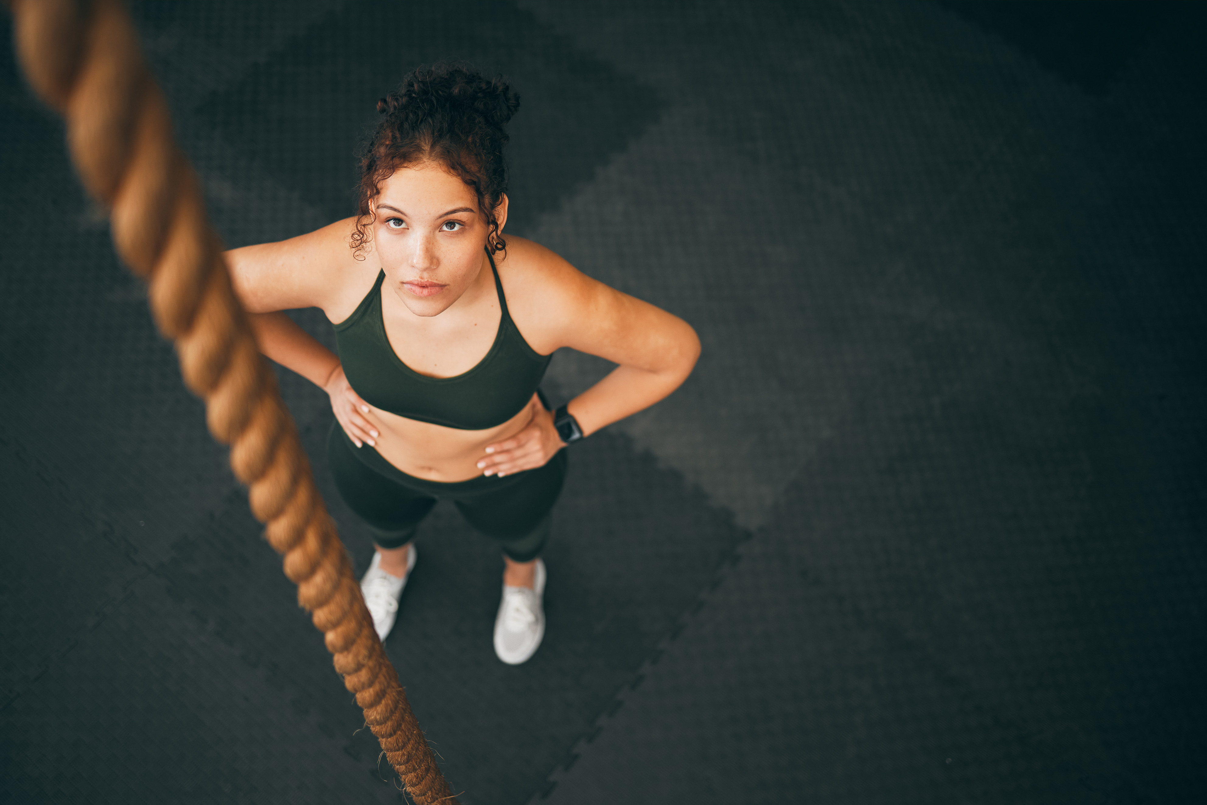 A woman in fitness clothing looks doubtfully up at a climbing rope