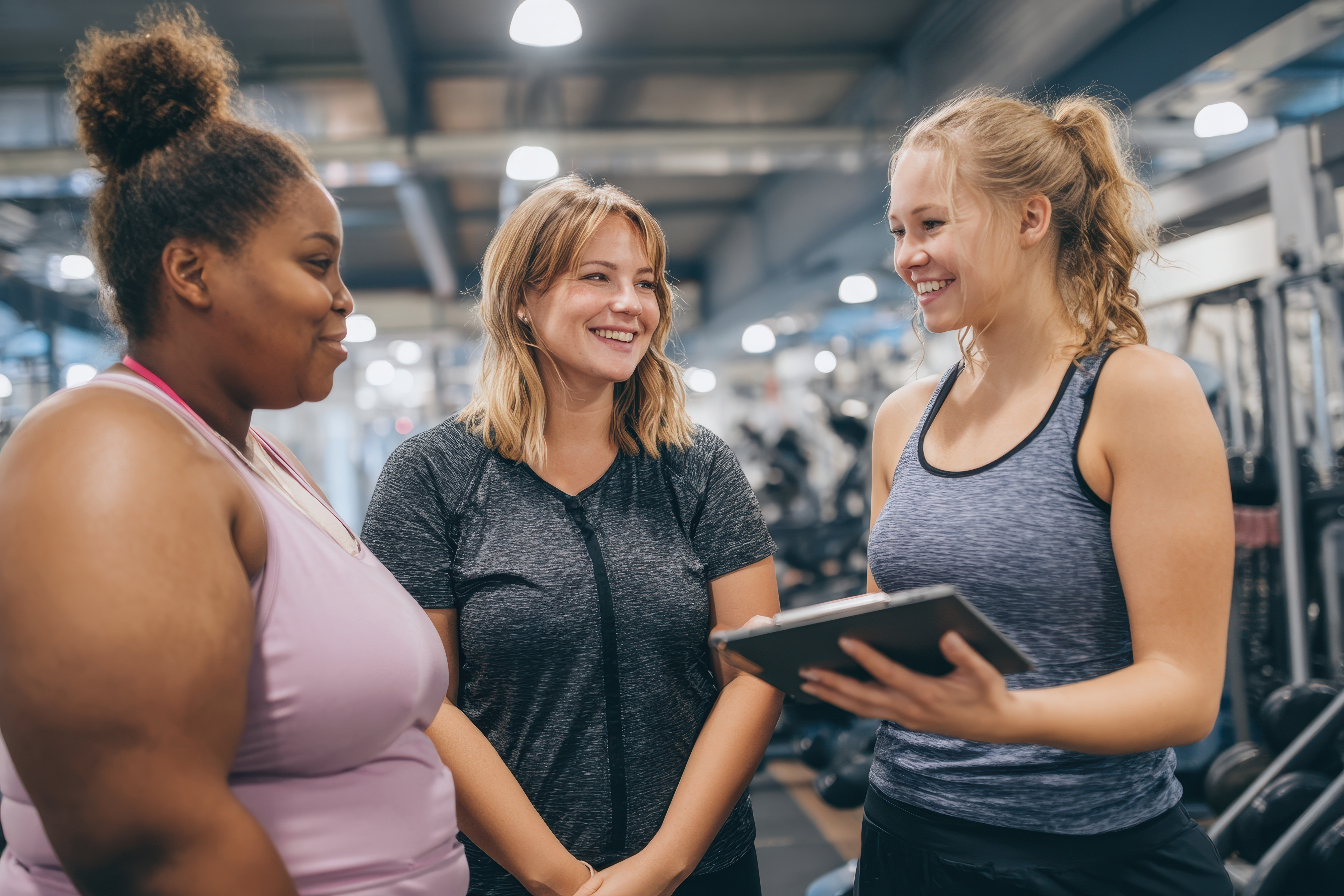 3 women in sports gear are chatting in a gym, 1 holds a tablet
