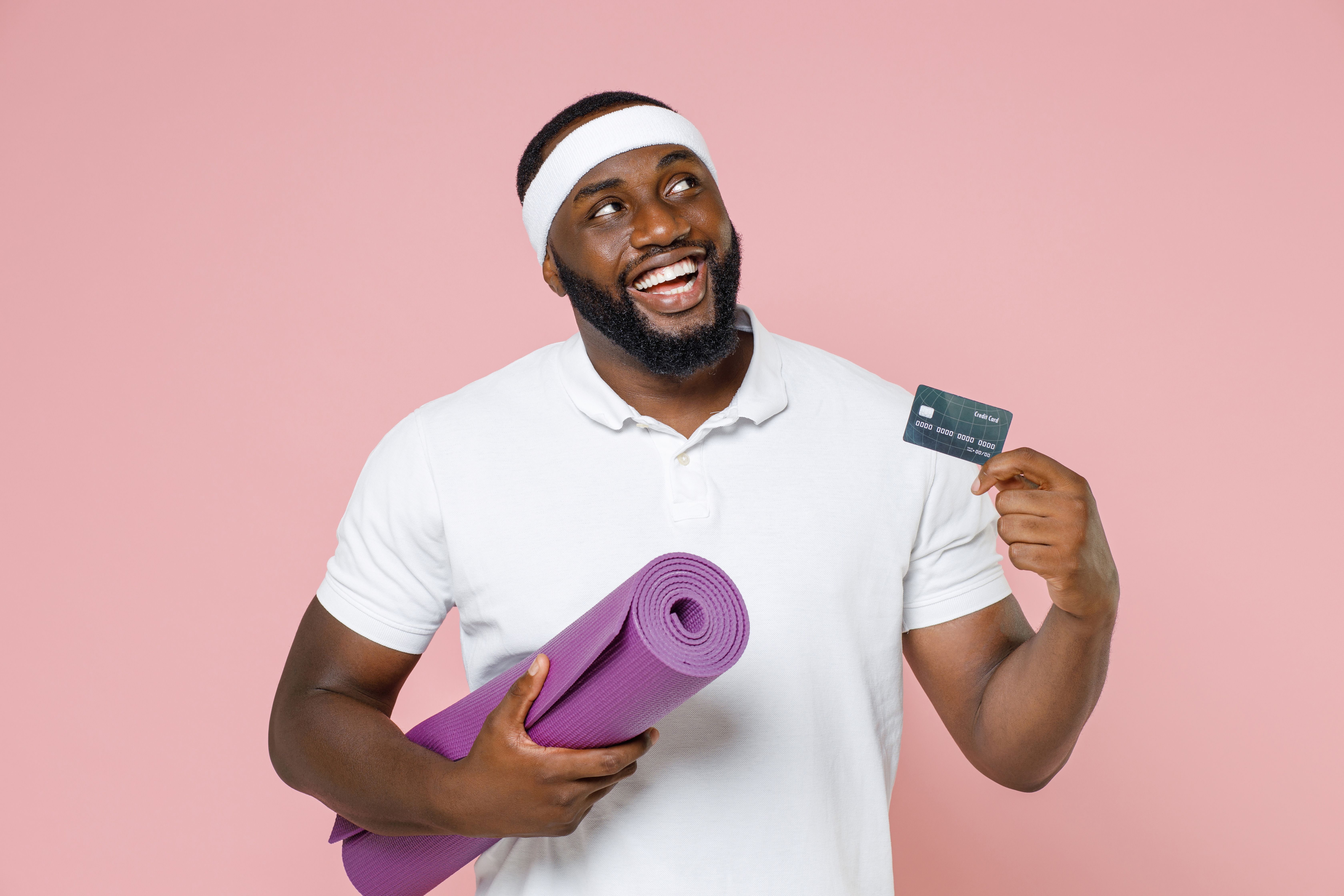 a man in a white polo shirt and headband looks up smiling whilst he holds a yoga mat in one hand and a bank card in the other