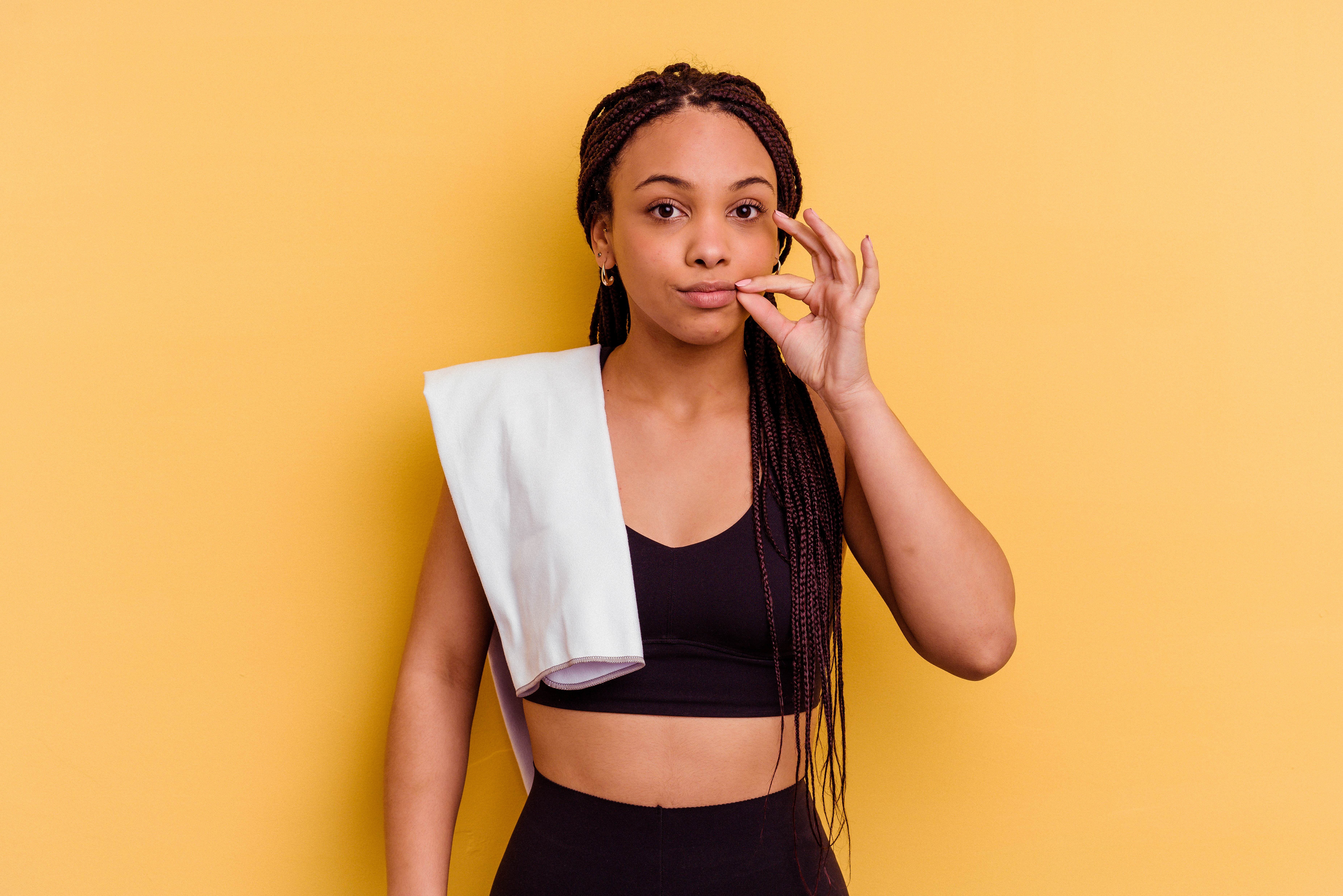 Young sport african american woman in gym gear with a towel over her shoulder isolated on yellow background with fingers on lips keeping quiet