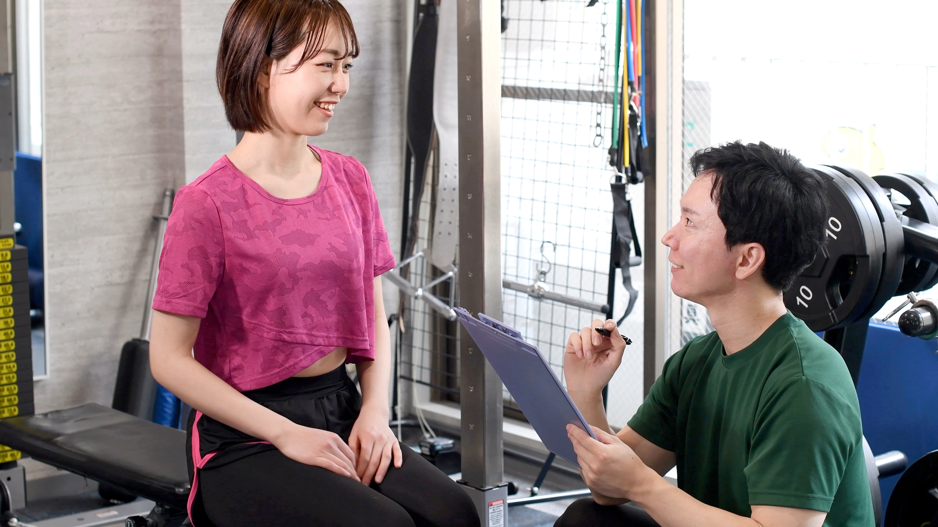 a man with a clipboard crouches next to a smiling woman sat on a gym bench.  They are in a gym and both wear gym clothing