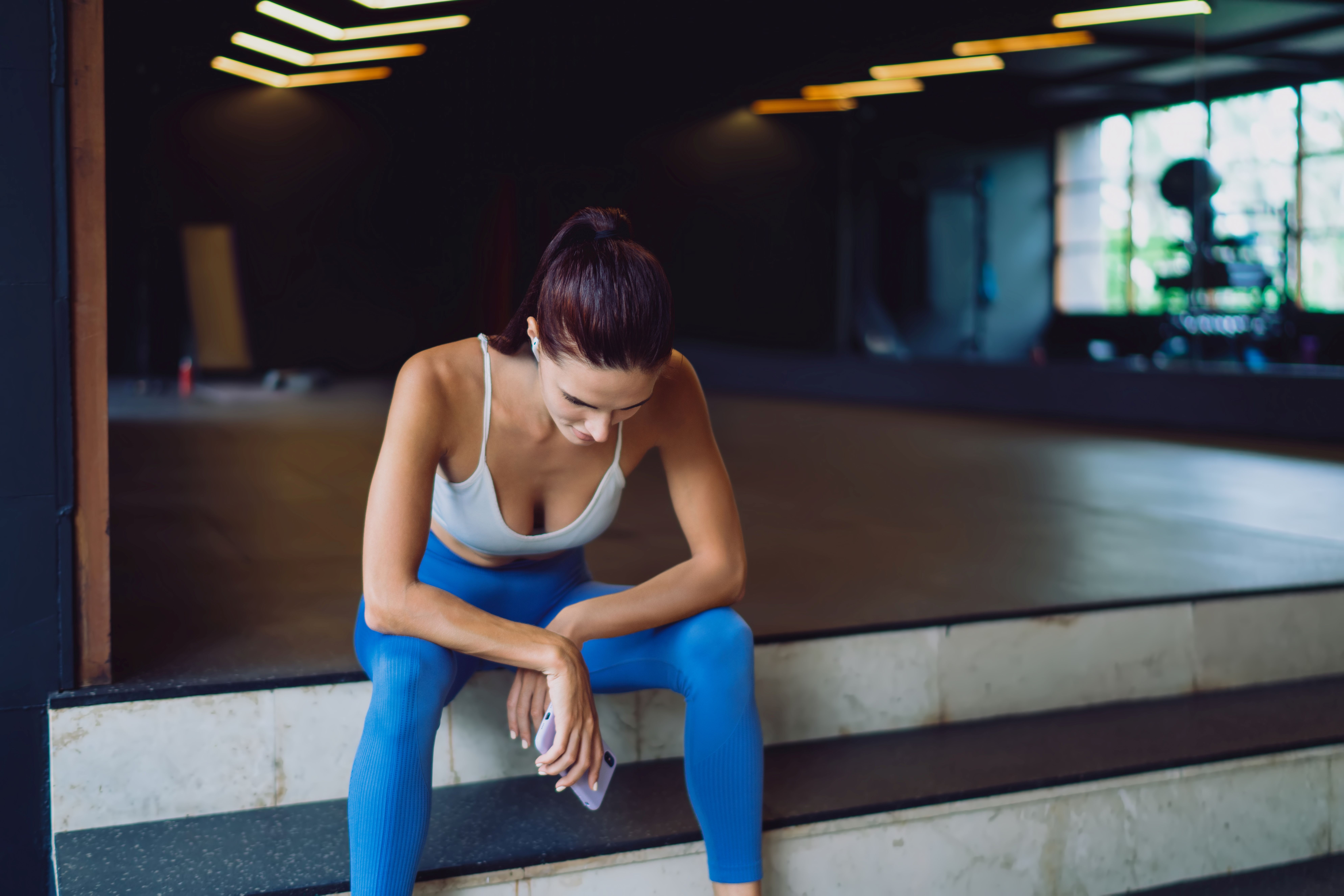 Tired young woman in sportswear sits on gym stairs, looking down 