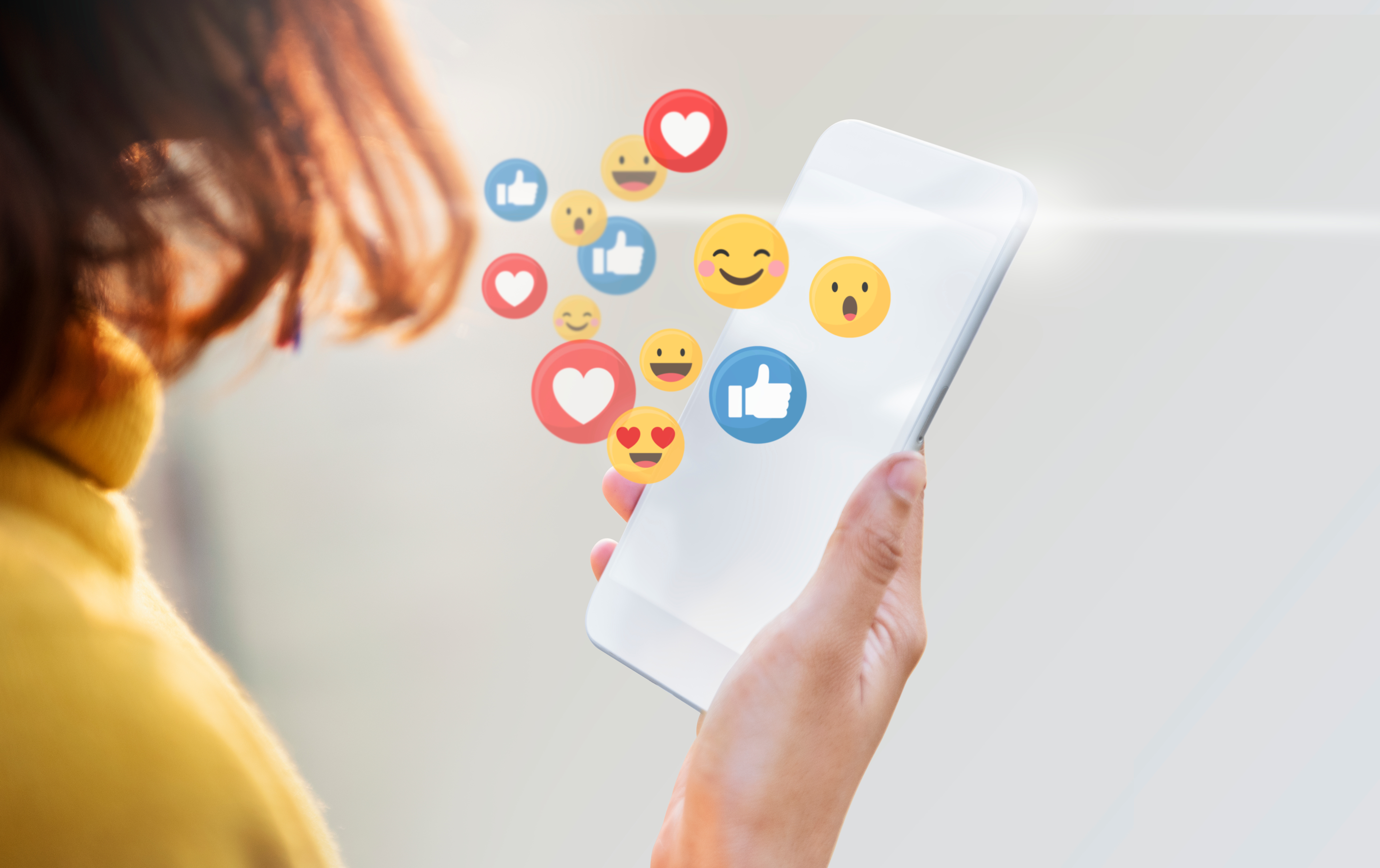 view over somone's shoulder as they look at a phone screen. hearts, likes and various smiley emojis hover over the phone screen
