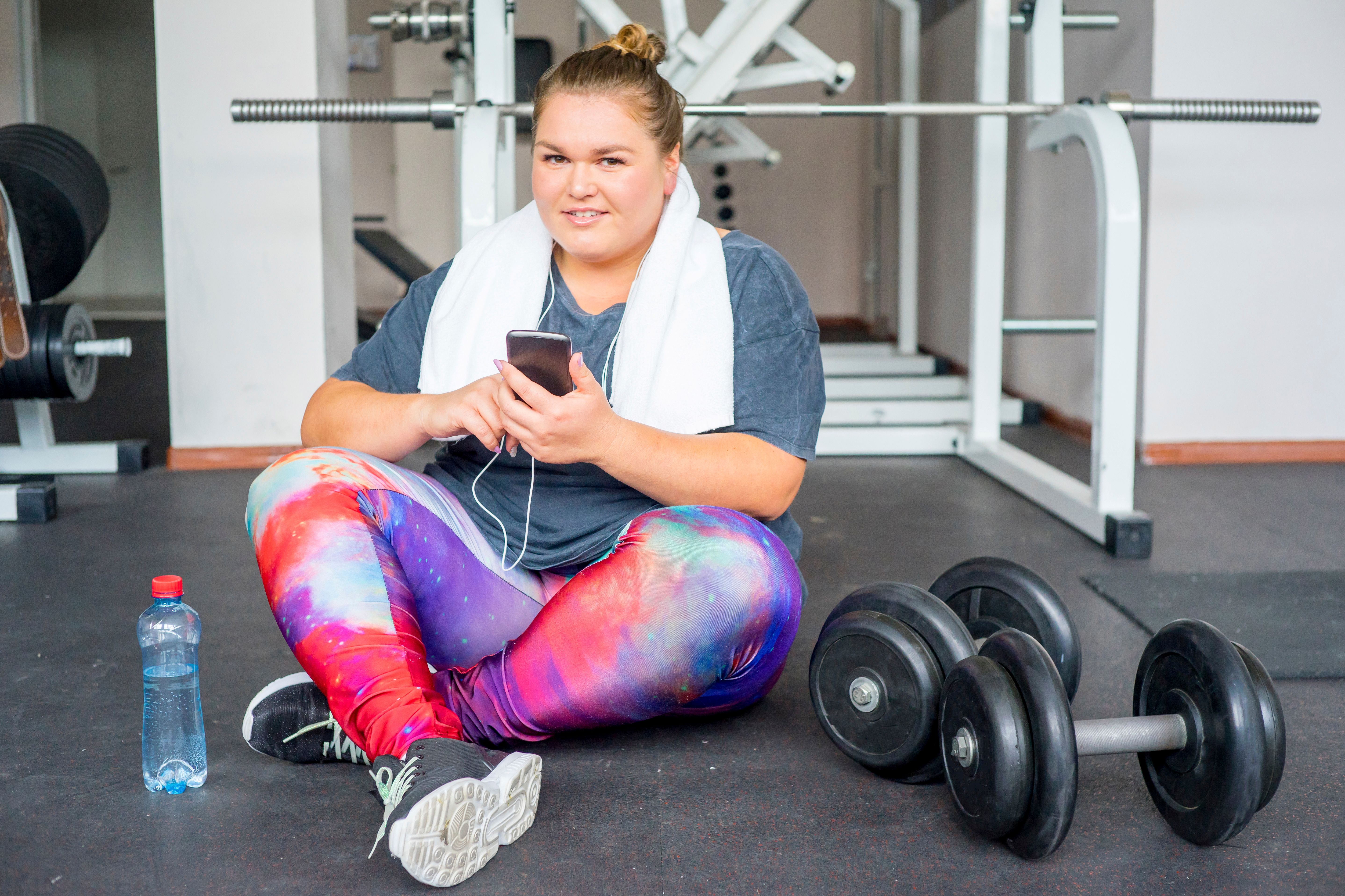 a woman in leggings and a t shirt sits on the floor in a gym with 2 heavy weights and a bottle of water next to her. She has a towel round her shoulders and her phone in her hands although she is looking at the camera