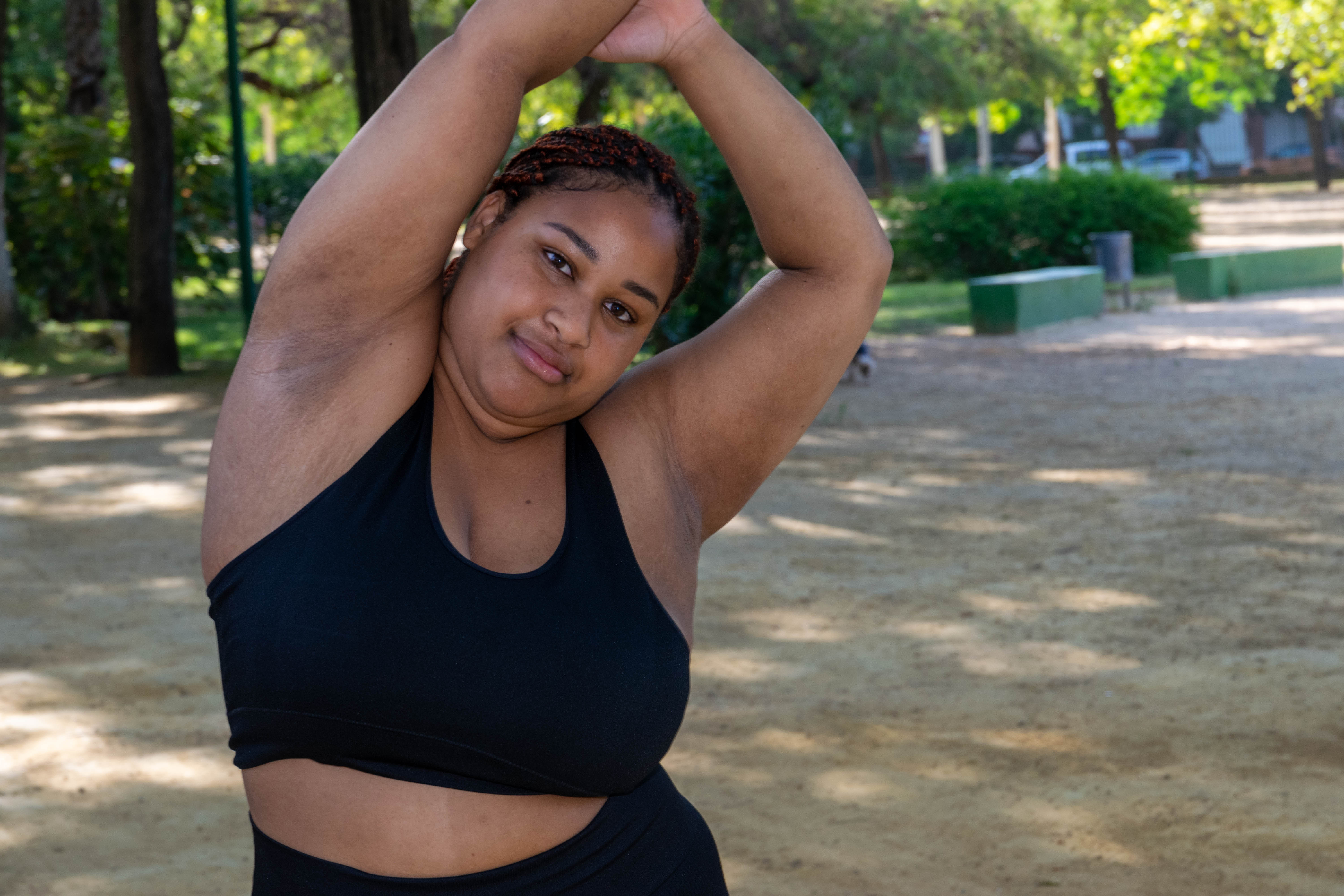 a larger dark skinned woman in a sports crop top and leggings is stretching in a park