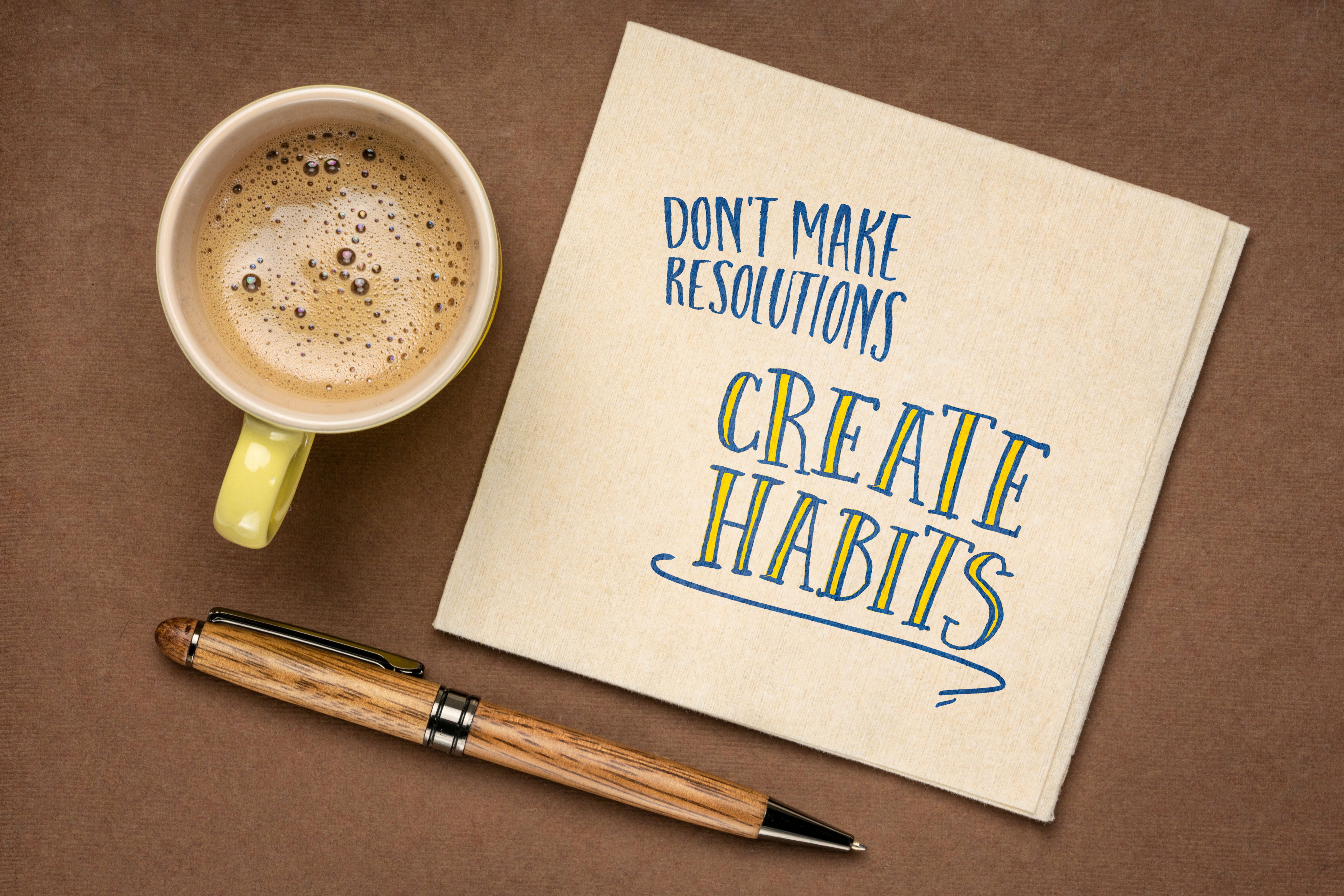 a napkin with the words do not make resolutions, create habits written on it lies on a table next to a cup of coffee and a pen
