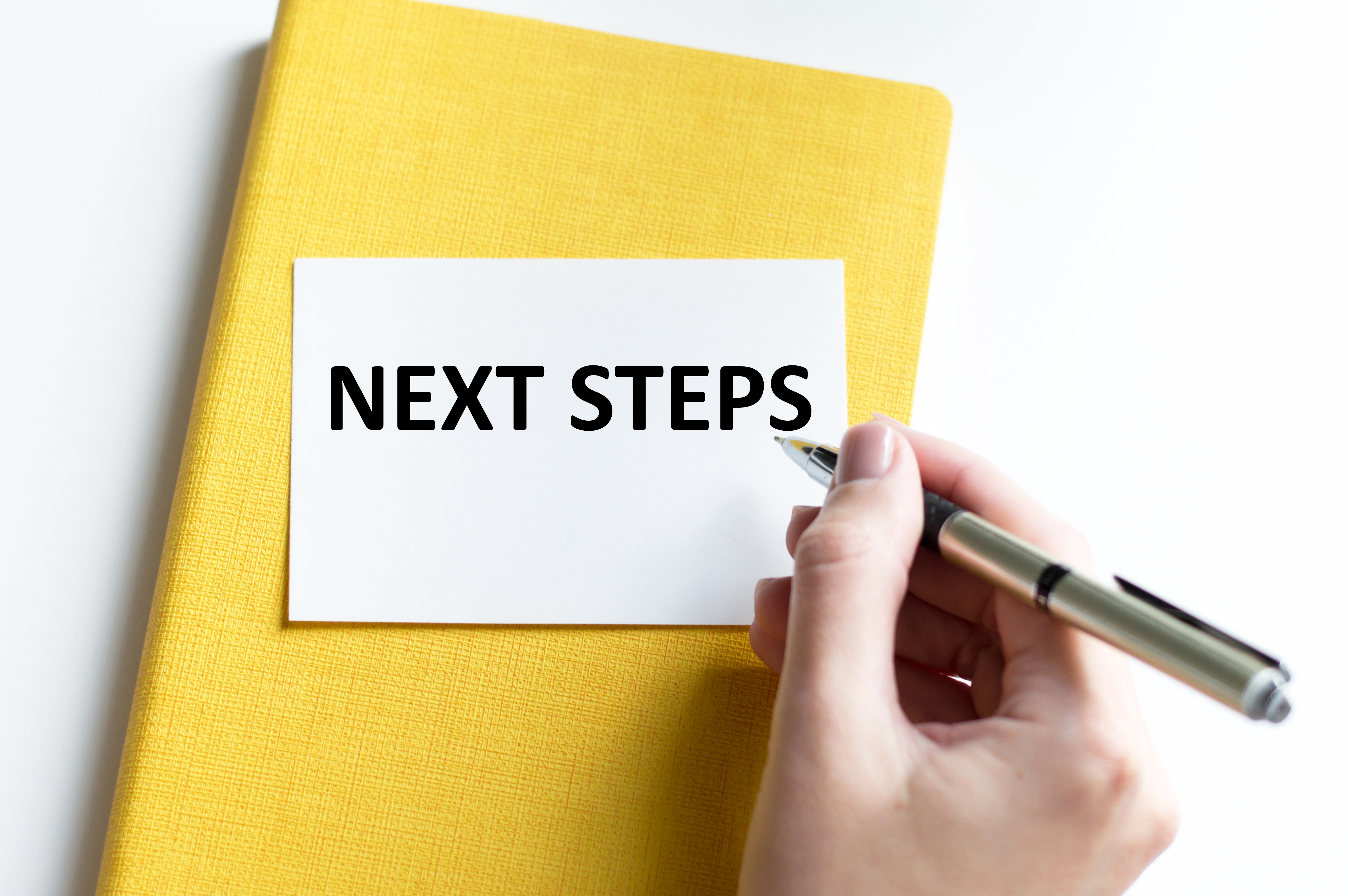 a piece of paper with the words next steps rests on a yellow covered book. A hand holding a pen rests next to it.