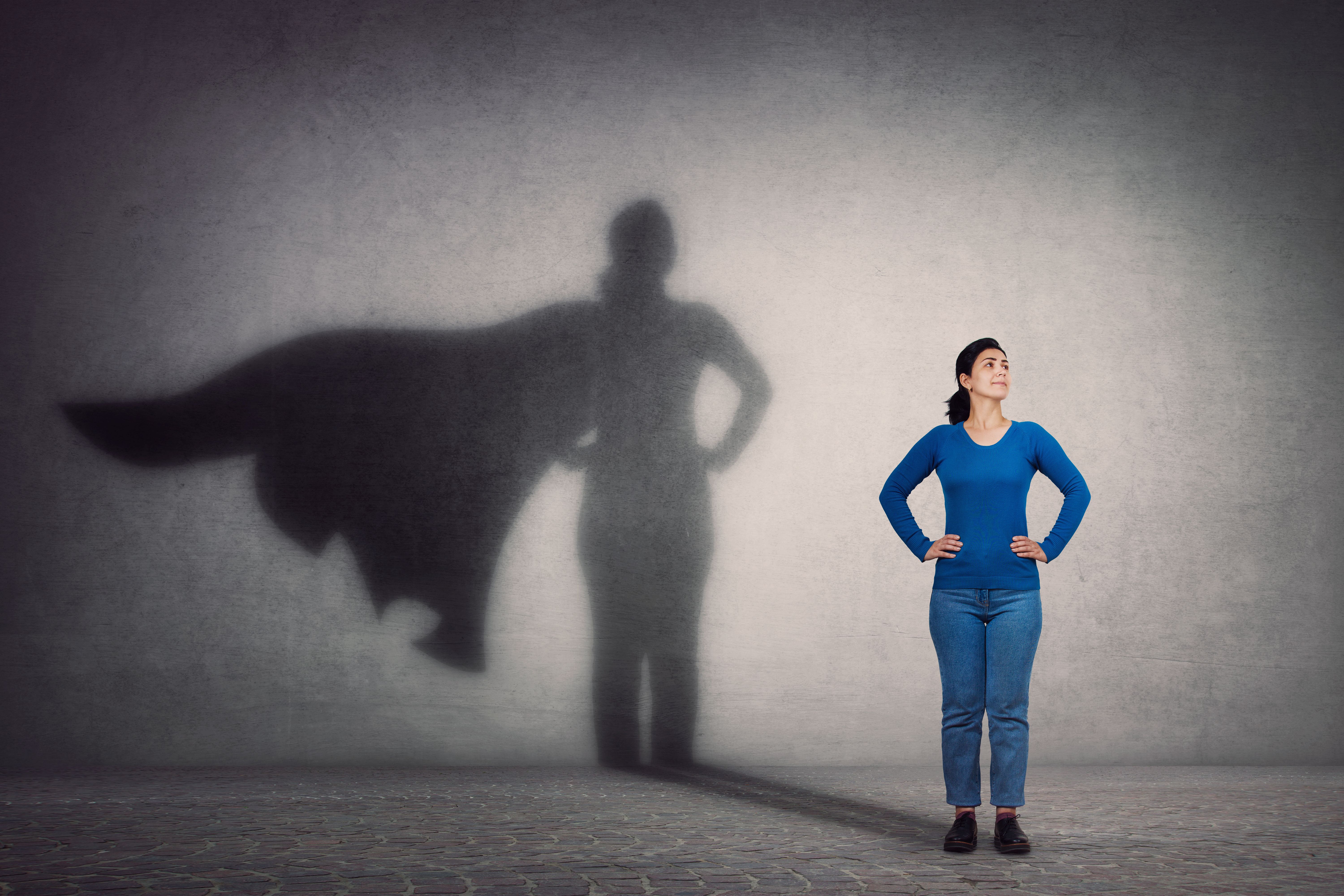 a smiling, confident woman stands with her hands on her hips casting a superhero with cape shadow on the wall behind her.