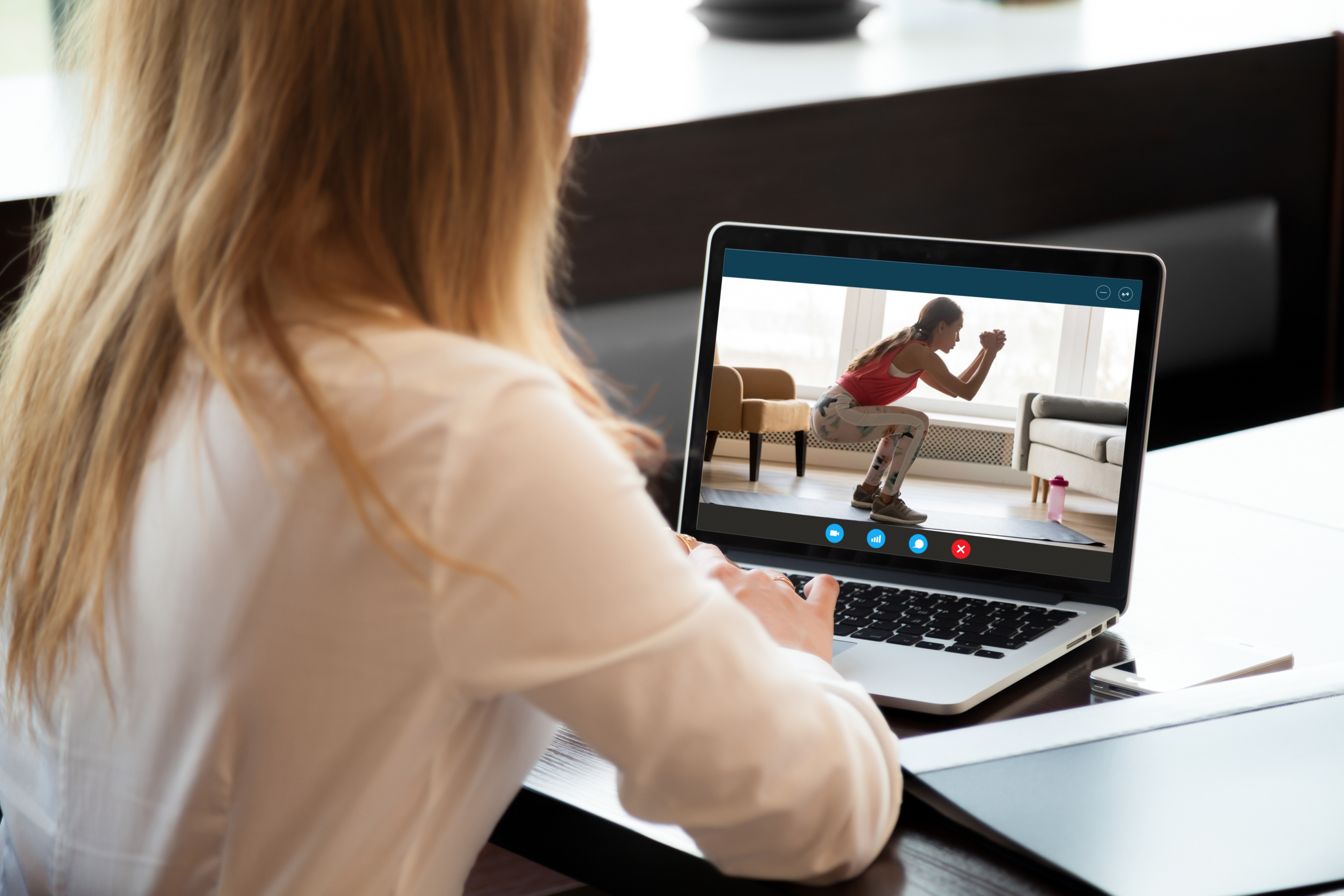 looking over the shoulder of a woman in a smart white blouse at the work she is doing on a laptop. On the laptop screen is an exercise video of a woman doing a squat.