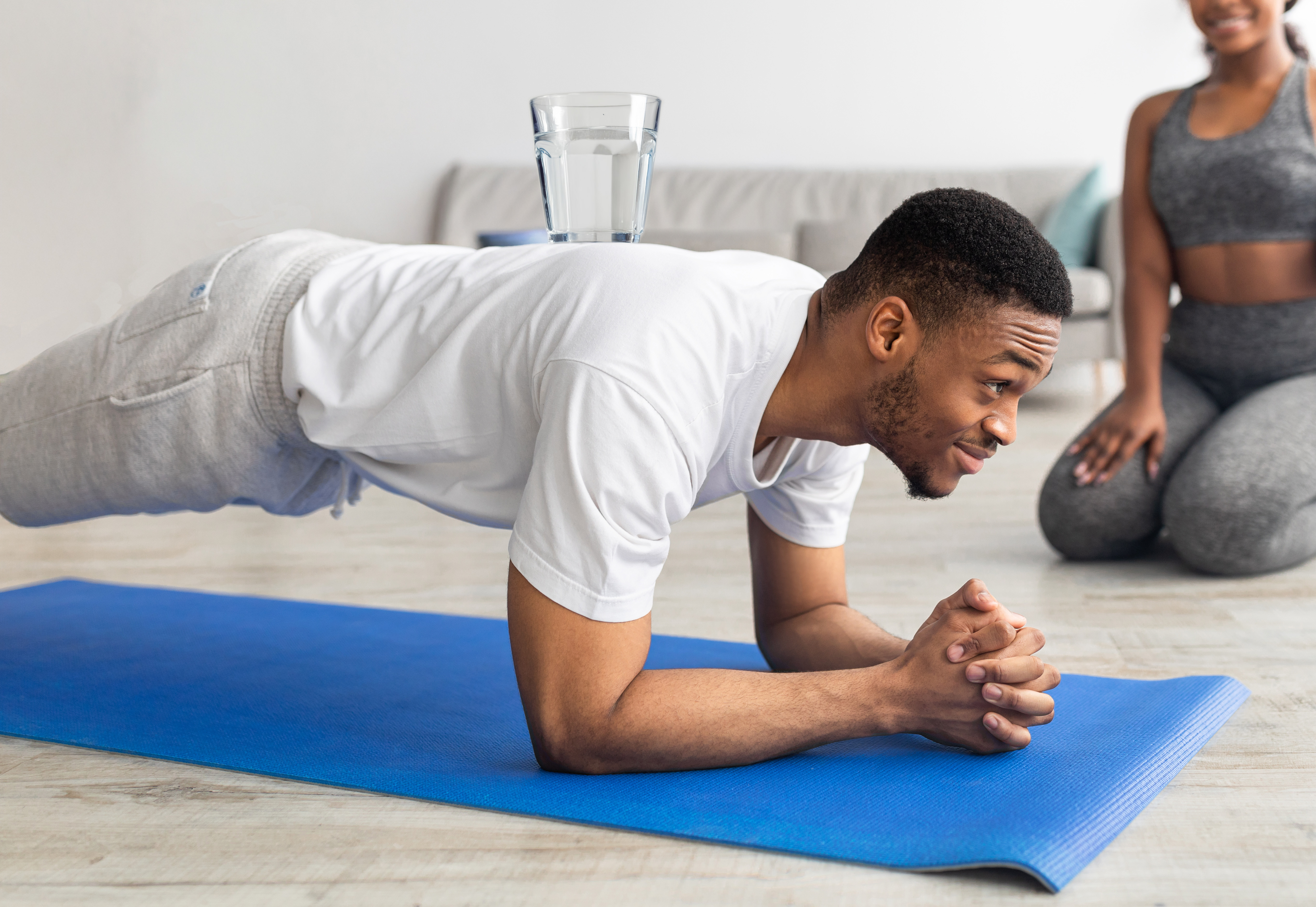 a man is in a lounge doing a plank with a glass of water on his back.  On the edge of the frame is a woman in gym kit watching on.