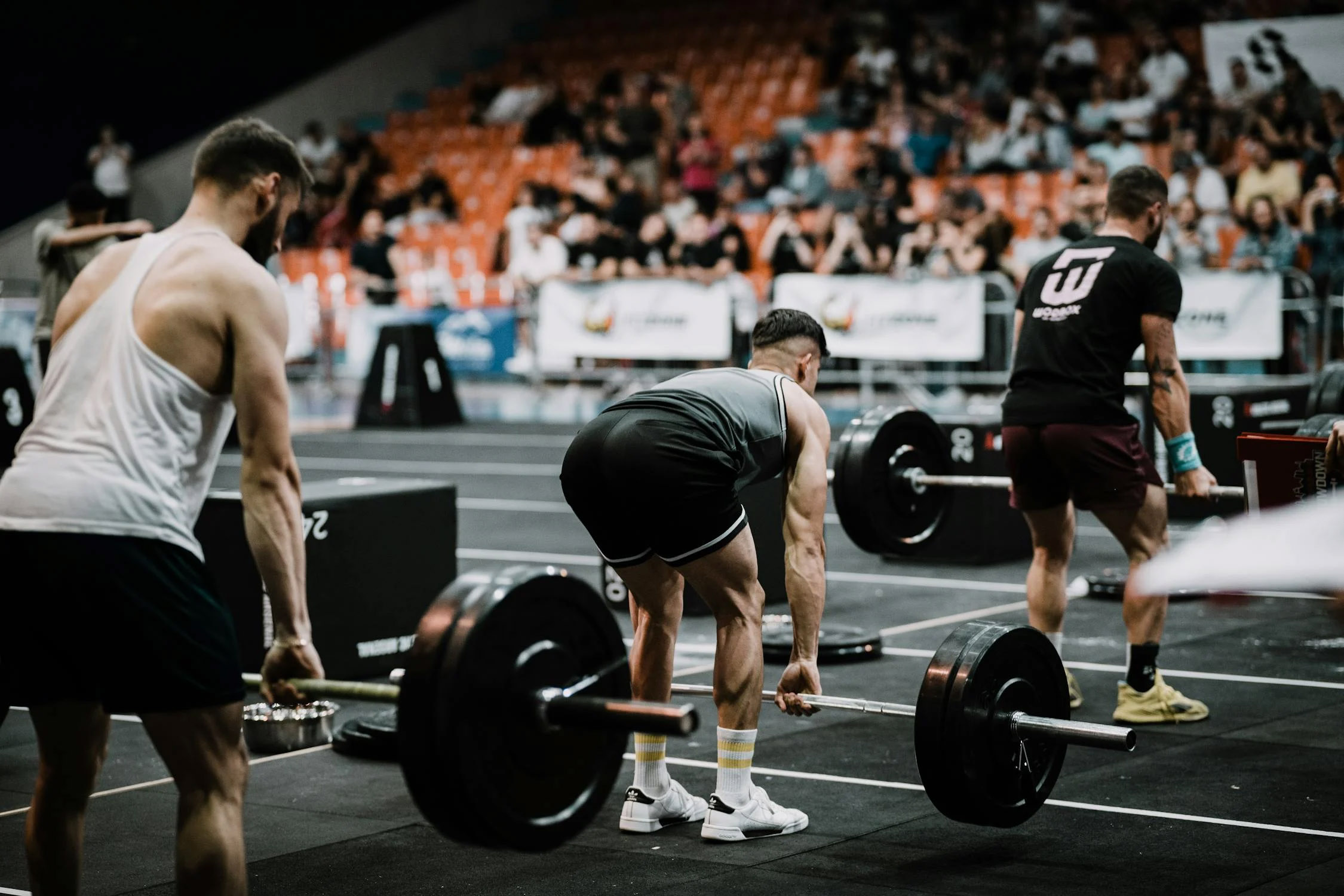 3 guys in shorts and t shirts are stood one behind the other deadlifting at a competition viewed dioagonally from the back