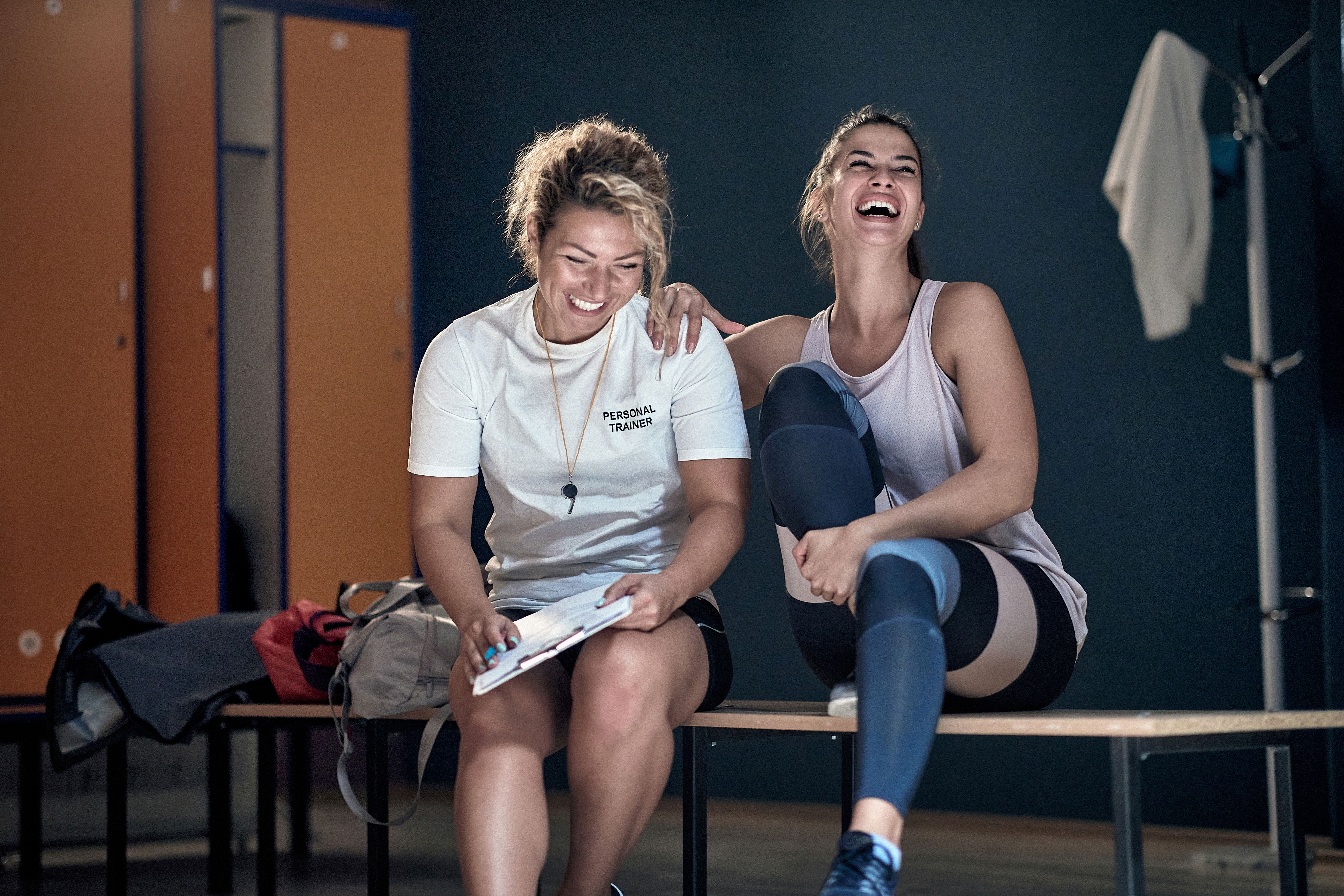 2 women in gym clothing sit on a bench in a changing room.  One leans on the others shoulder and they are both laughing