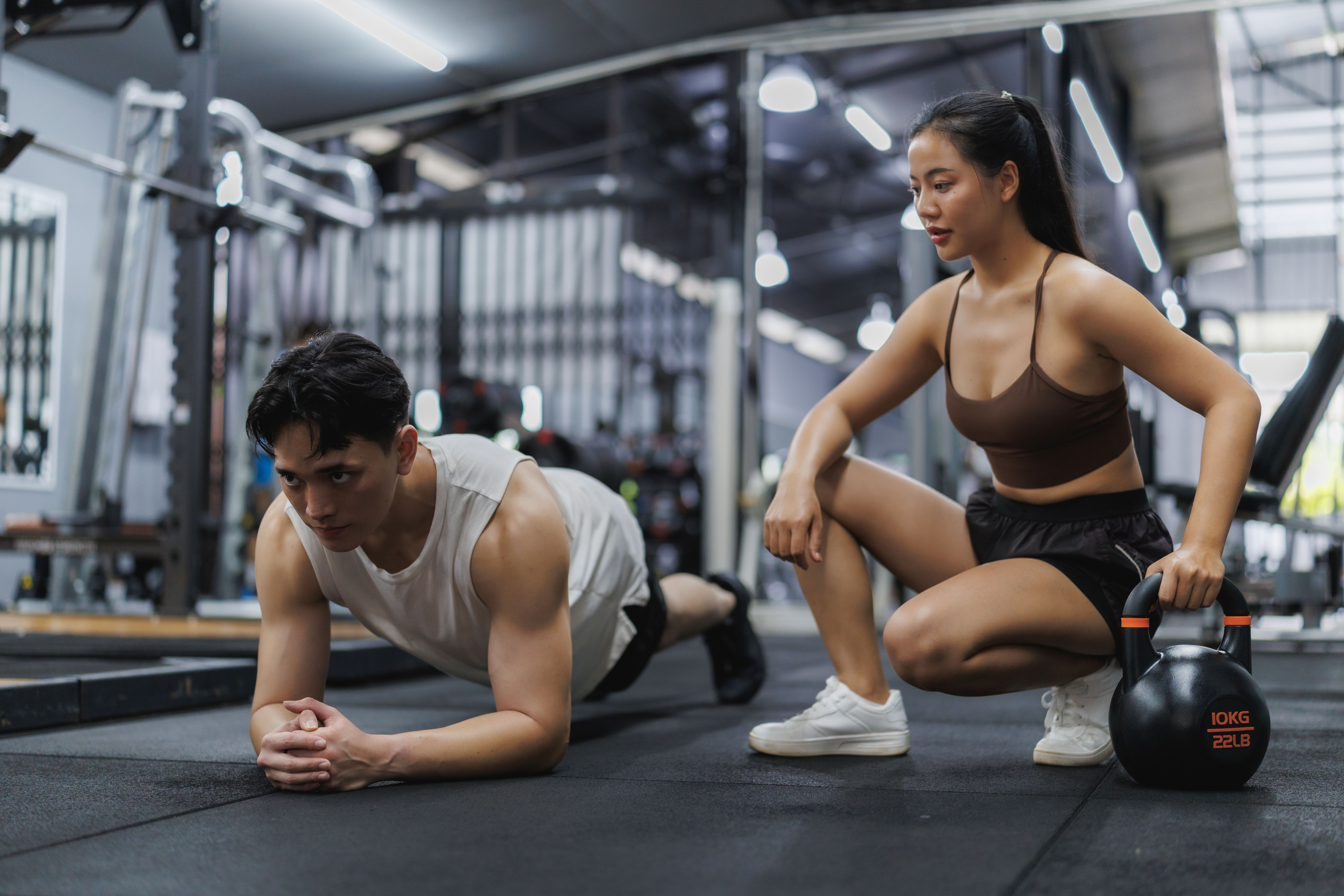a female trainer is crouched with one hand on a kettle bell watching a man do a plank next to her