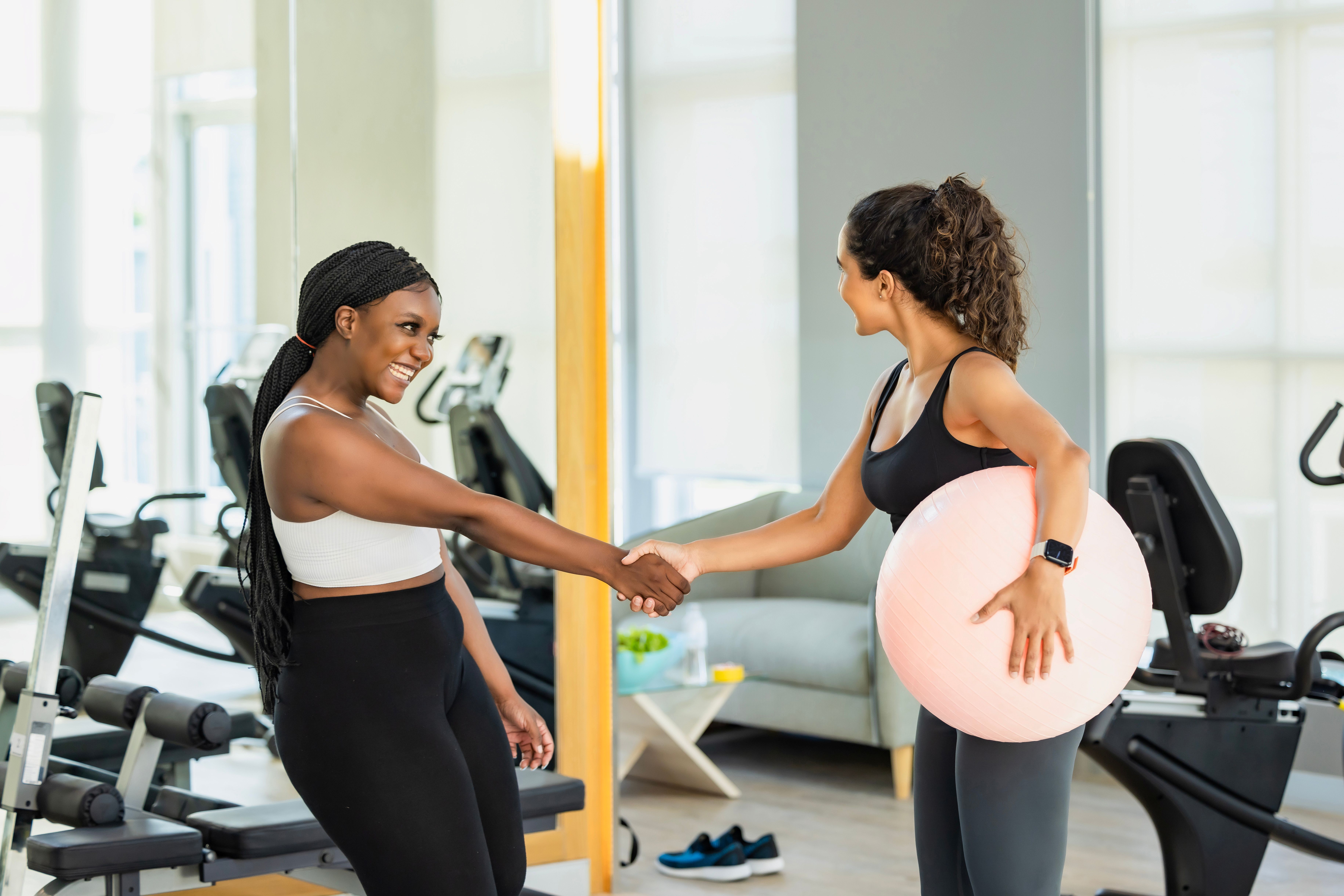 a woman holding a fitball under one arm shakes the hand of another lady.  Both wear fitness gear and in the background there are gym machines as well as an armchair and coffee table