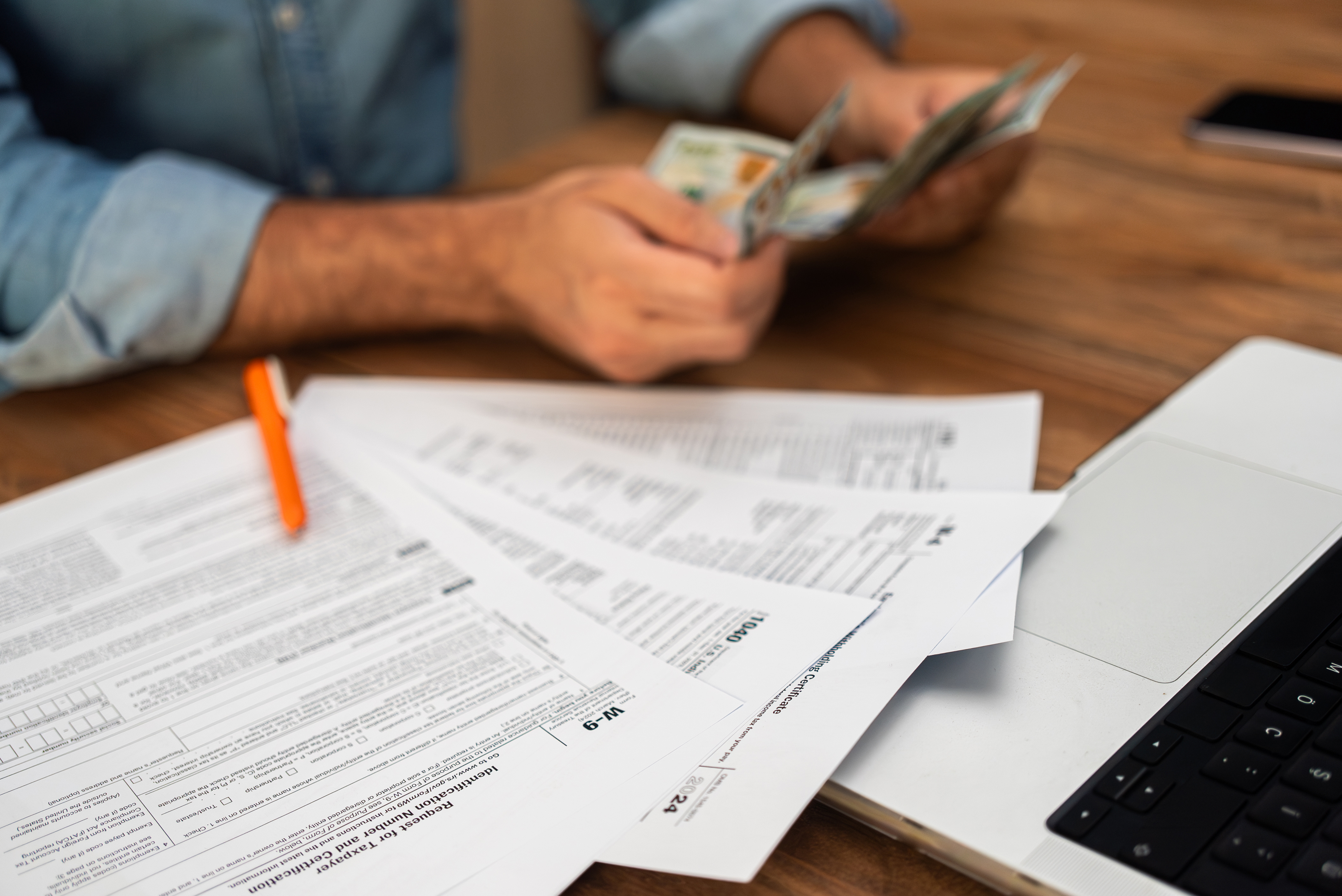 Man in denim shirt counting cash while examining tax forms on a wooden desk.