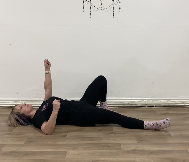a woman lies on her back on the floor with her left arm pointing up towards the ceiling and left leg bent at the knee with the foot firmly on the floor