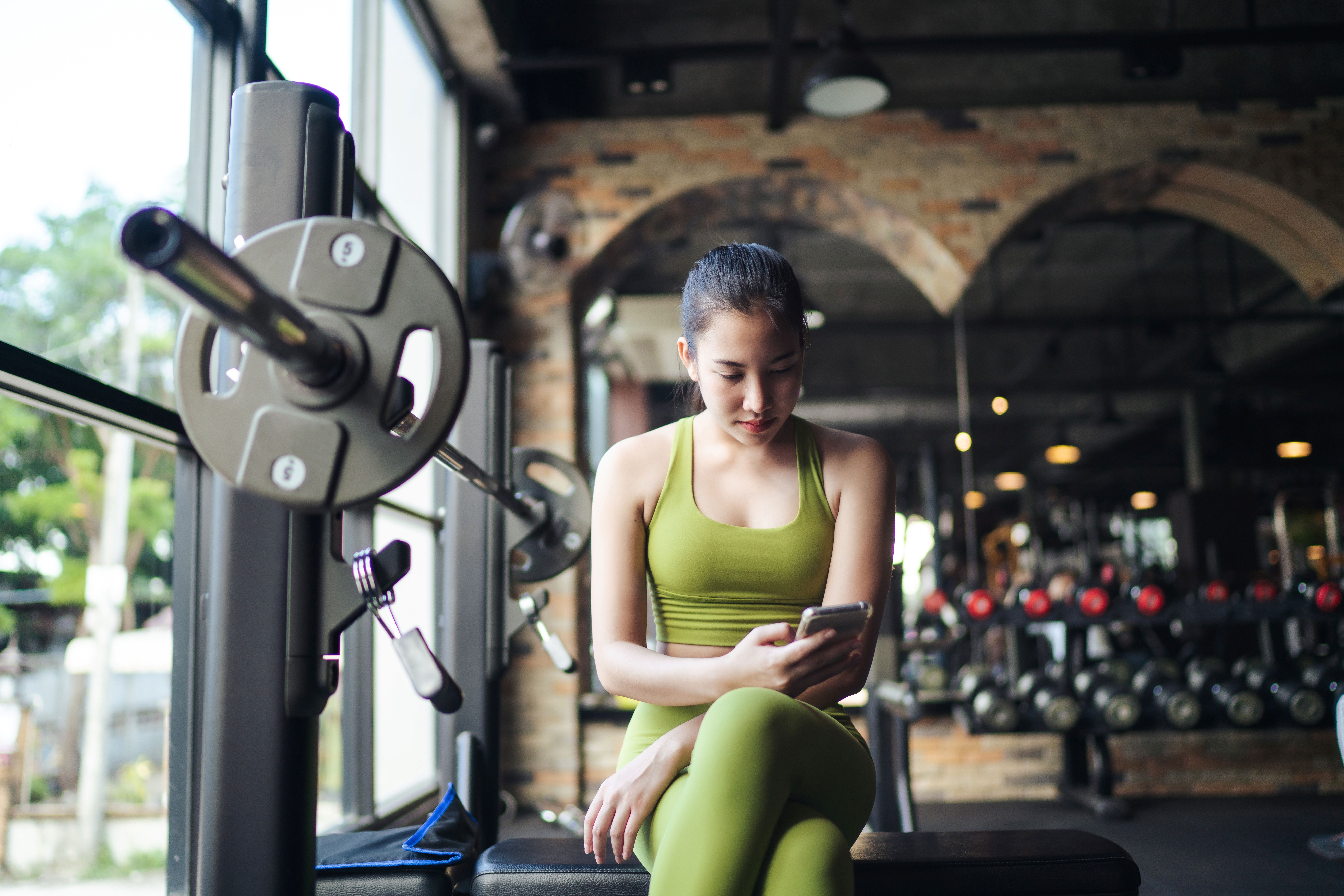 a young woman wearing fitness clothing is sat on a bench in a gym looking at her phone