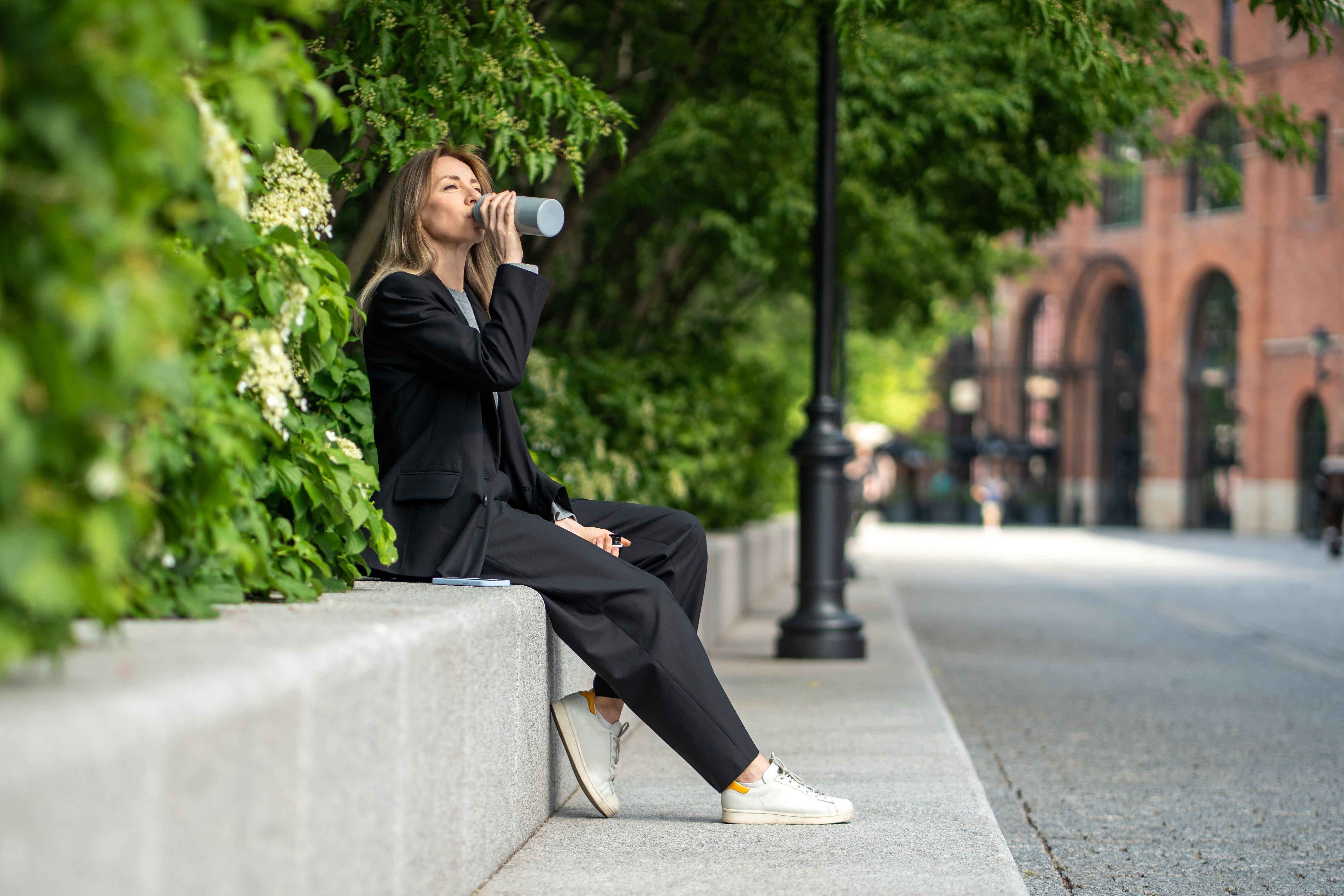 a woman in a black track suit is drinking from a water bottle while sitting on a low wall in front of some trees.  