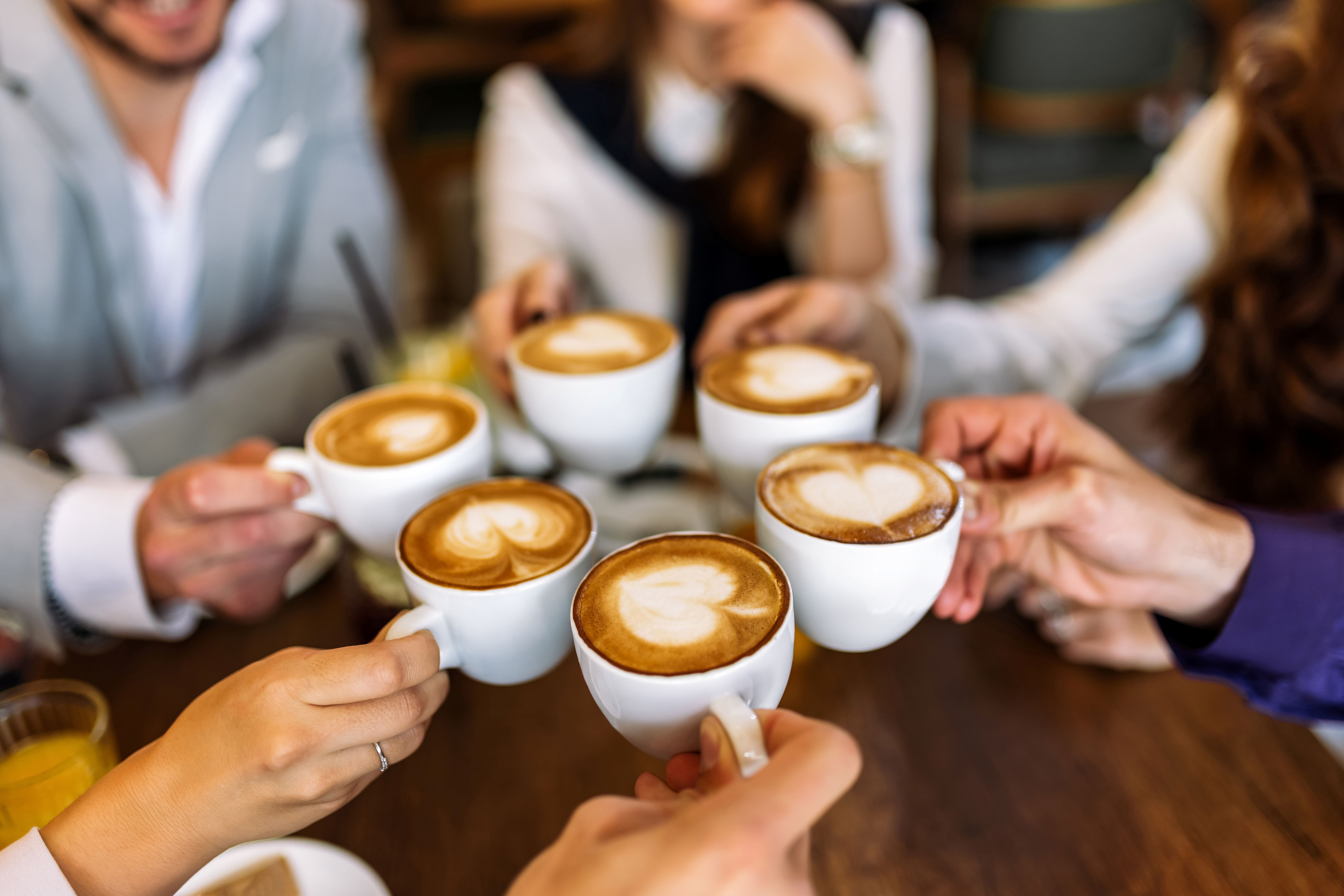 6 people each hold a cup of coffee in a circle.  Only the coffees and hands are in focus with the individuals sat round the table blurred