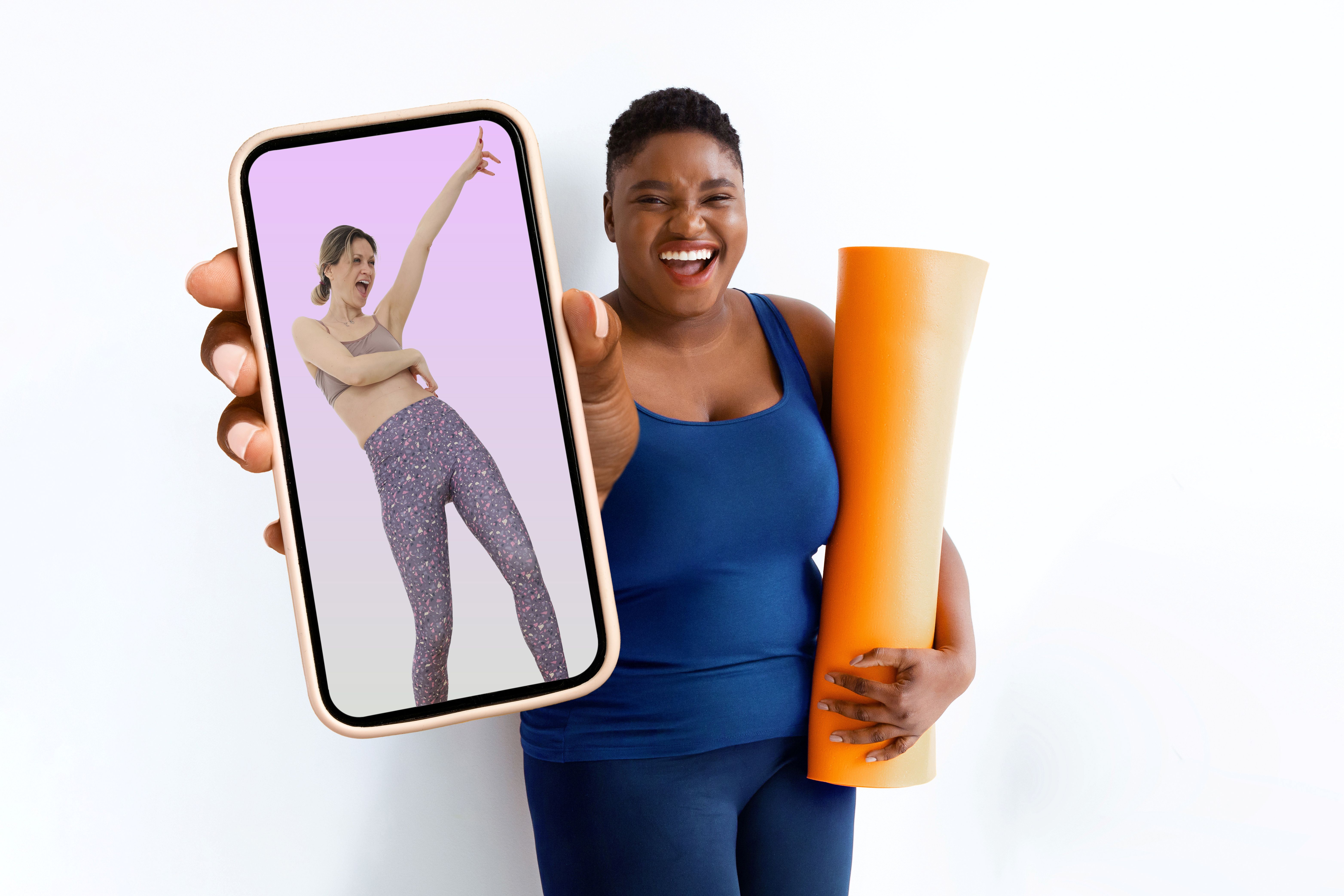a woman in gym gear holds an exercise mat and has a big smile on her face as she holds up a mobile phone showing a different woman in leggings who looks like she's dancing