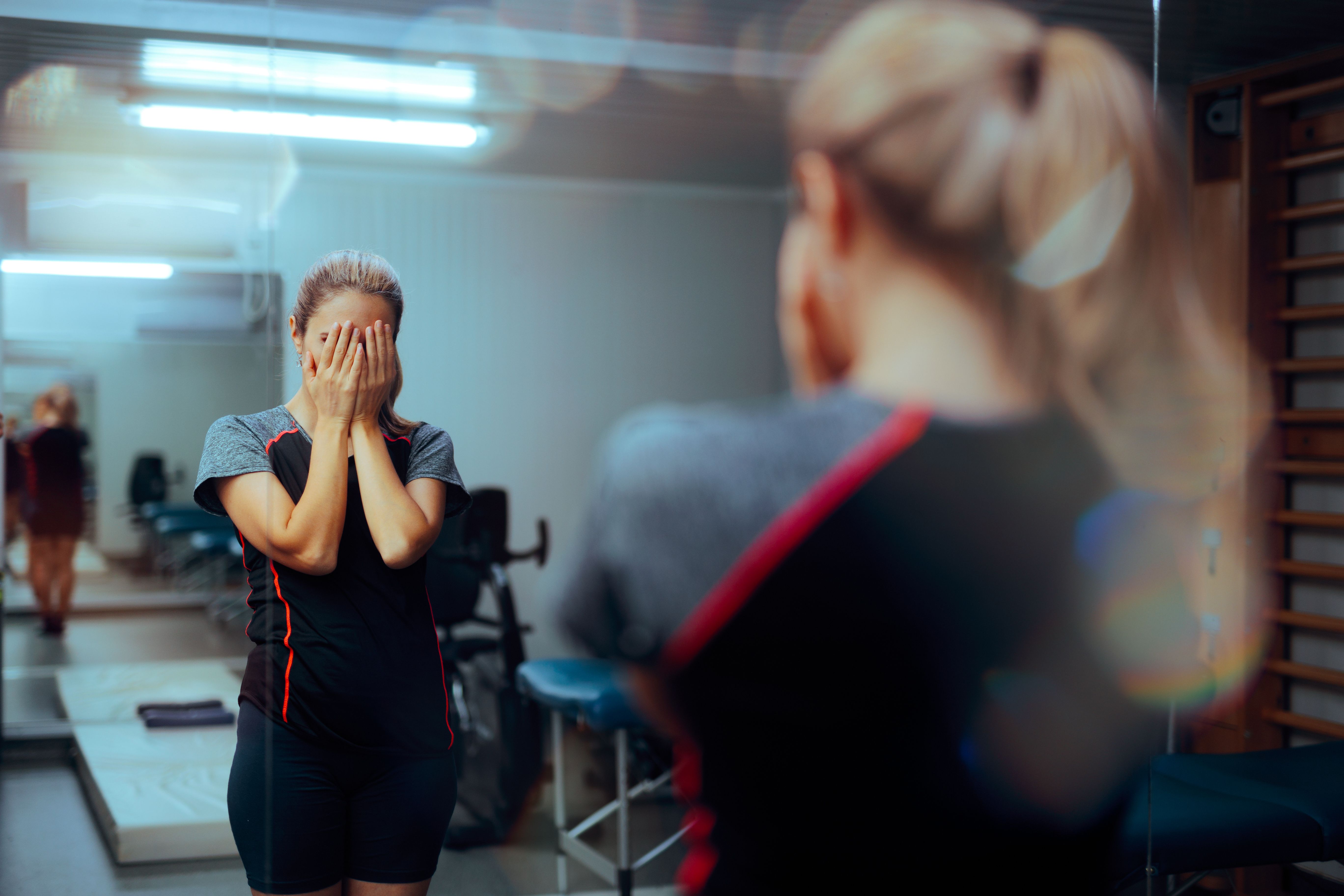 a woman in gym clothing stands in front of a mirror in the gym with her hands over her face in distress