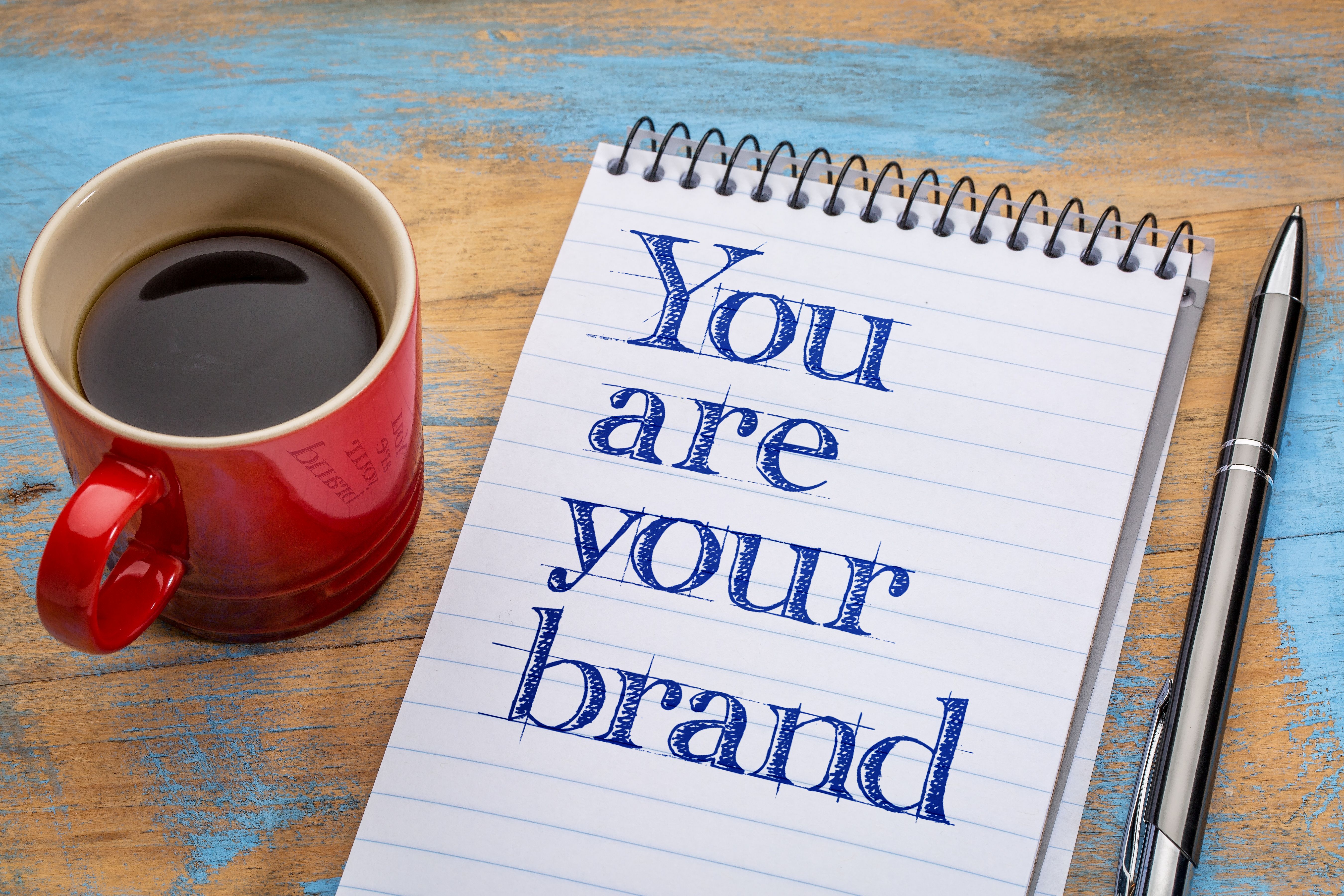 a mug of coffee sits next to a note pad and pen. The words You Are Your Brand are written on the notepad